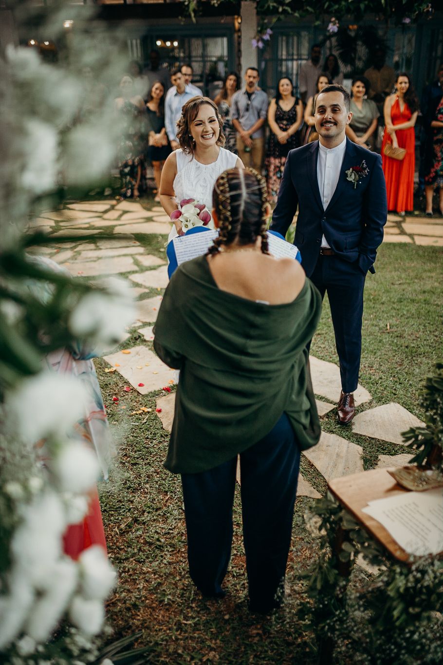 Wedding guests in formal attire stand on a lawn during an outdoor ceremony.