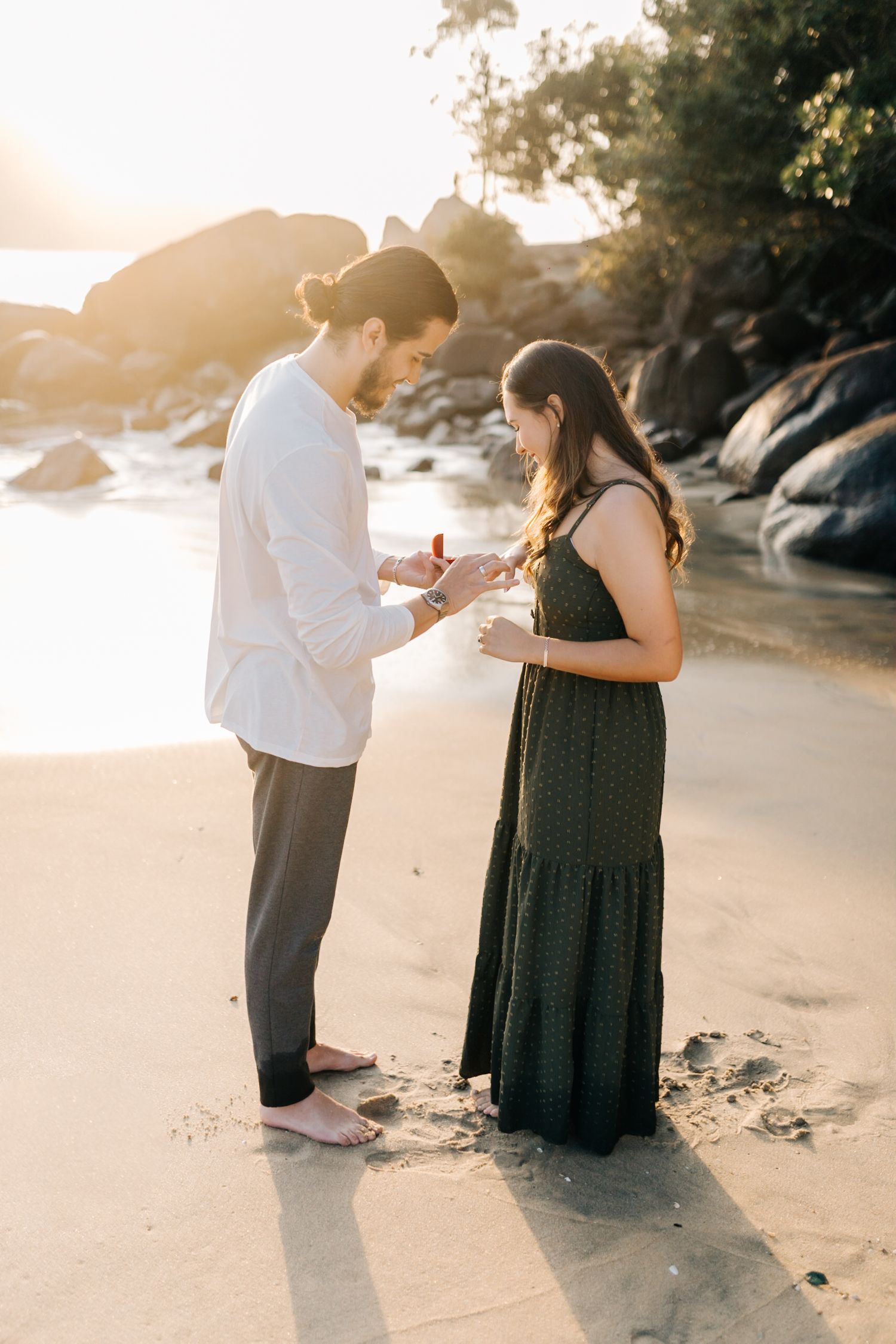 Couple standing together on beach sand near rocky cliffs at sunset.