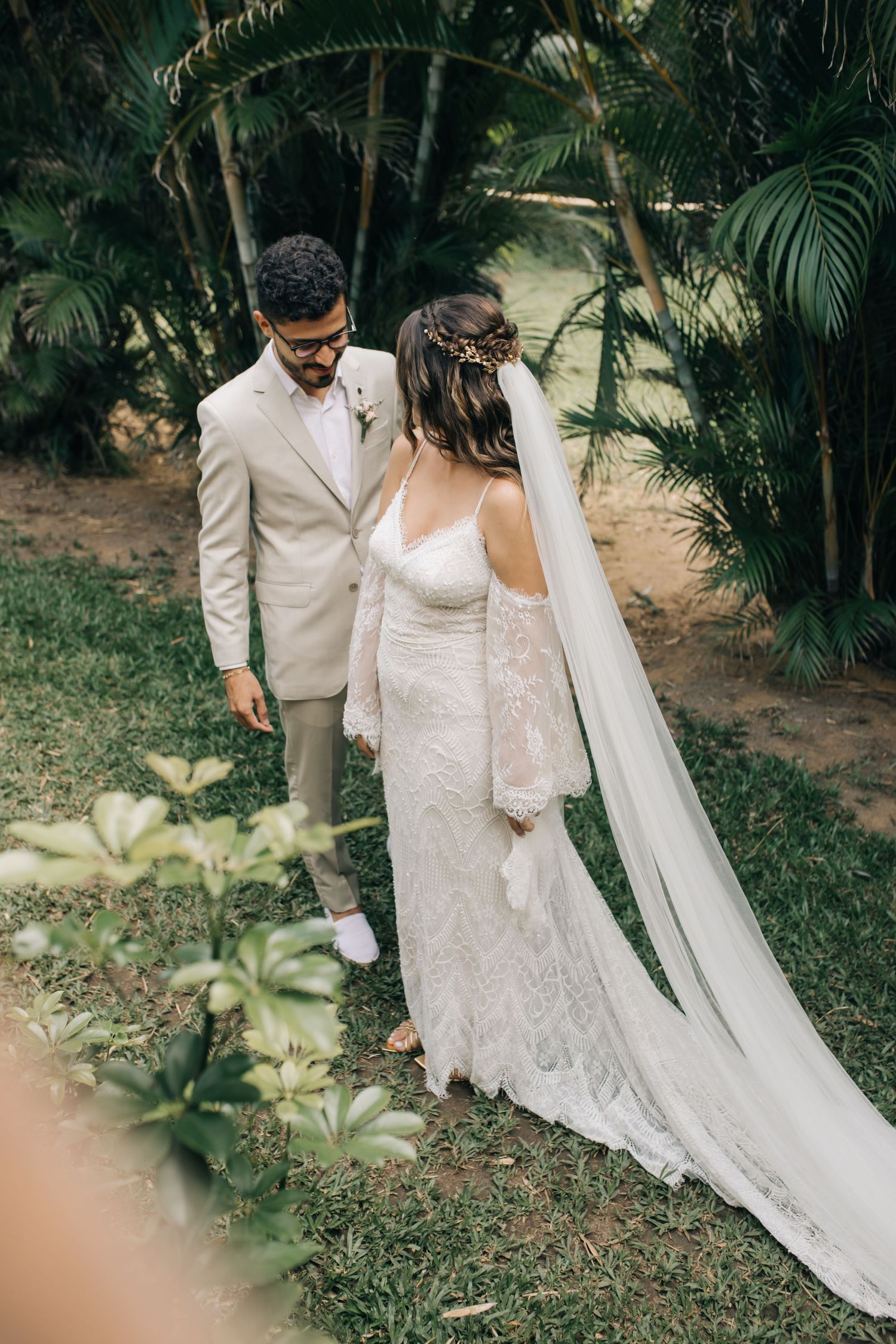 A couple in wedding attire poses in an elegant garden setting with palm trees and lush greenery.