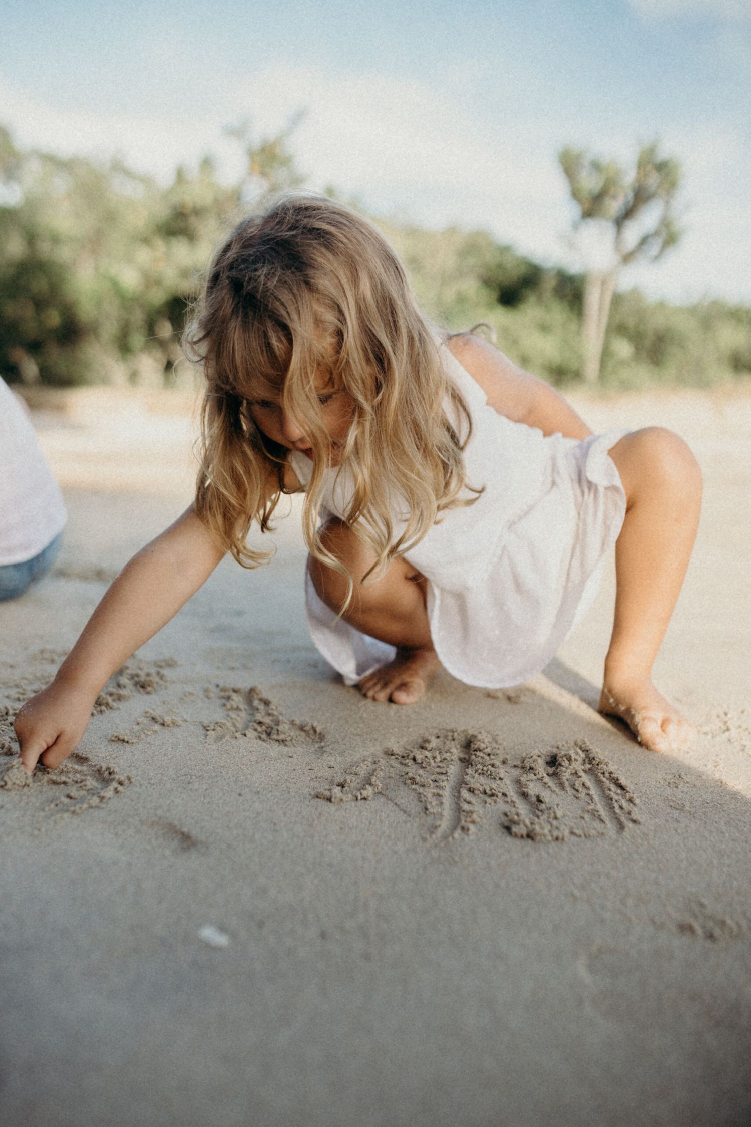 Child in white dress playing and crawling in beach sand during daytime.
