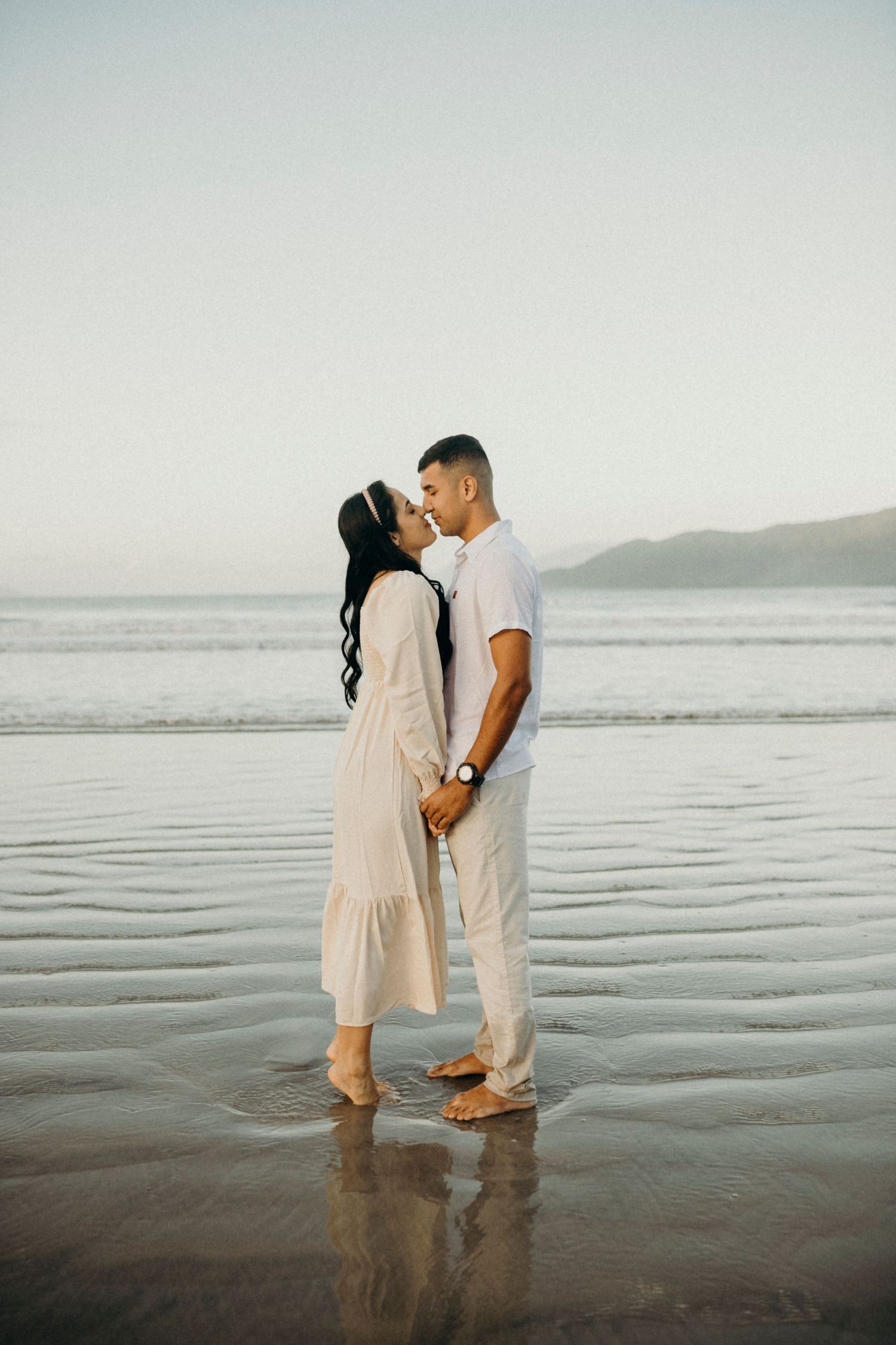 Two people in coordinated white outfits stand close together on wet beach sand at dusk.