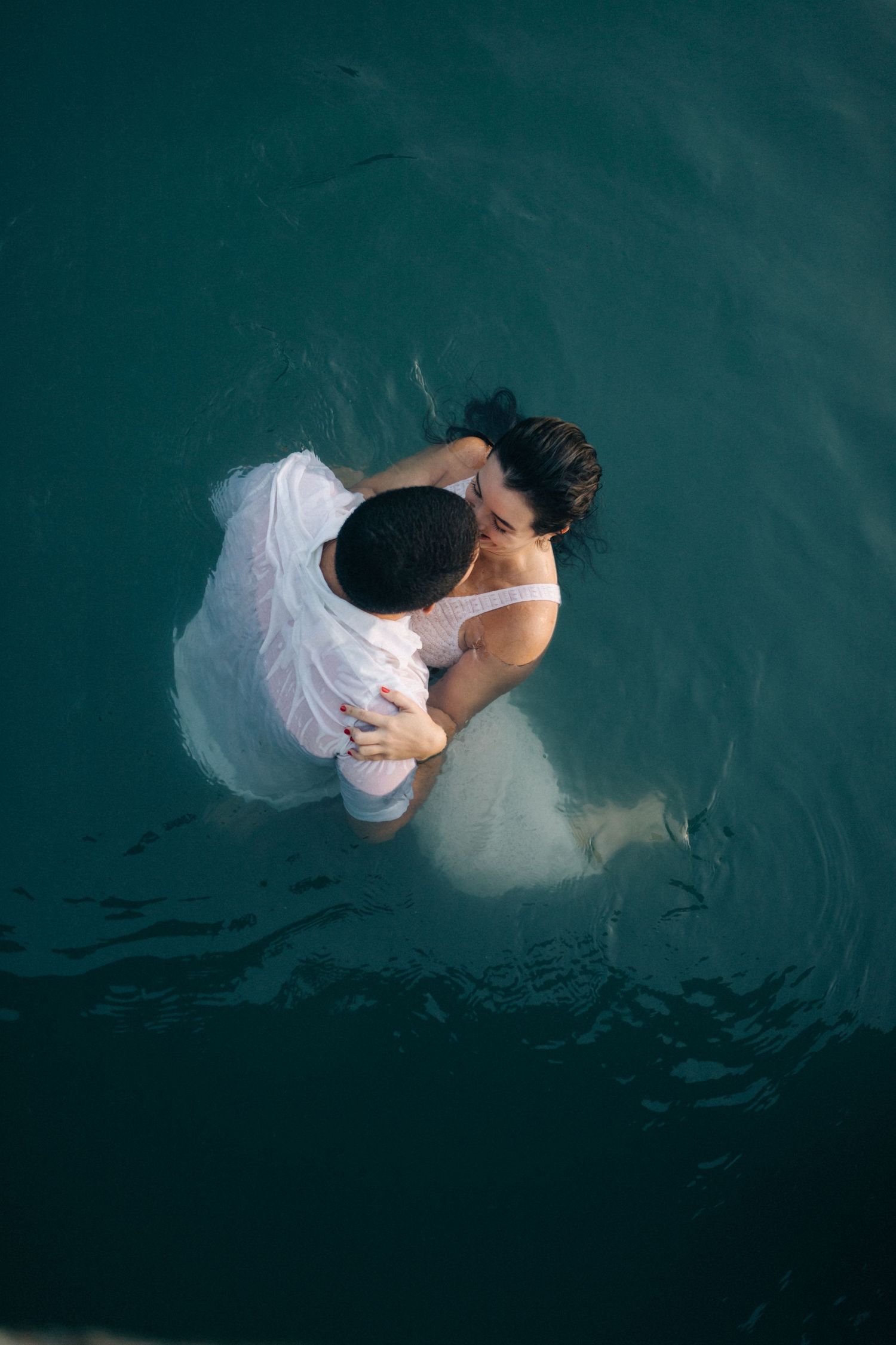 A couple embraces while floating in deep turquoise water, viewed from above.