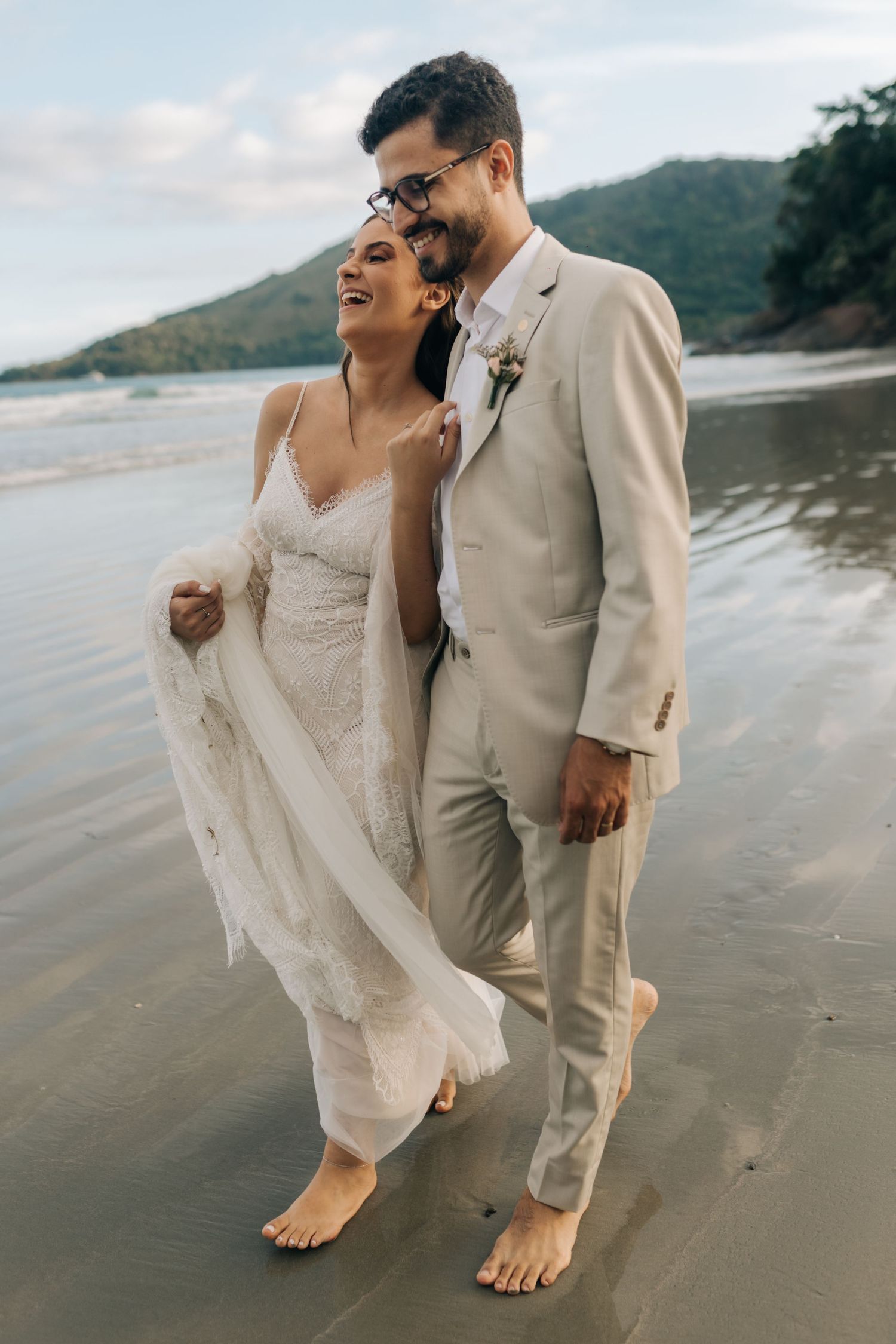 A romantic beach wedding photo features a couple in white and beige attire laughing together barefoot on the sand.