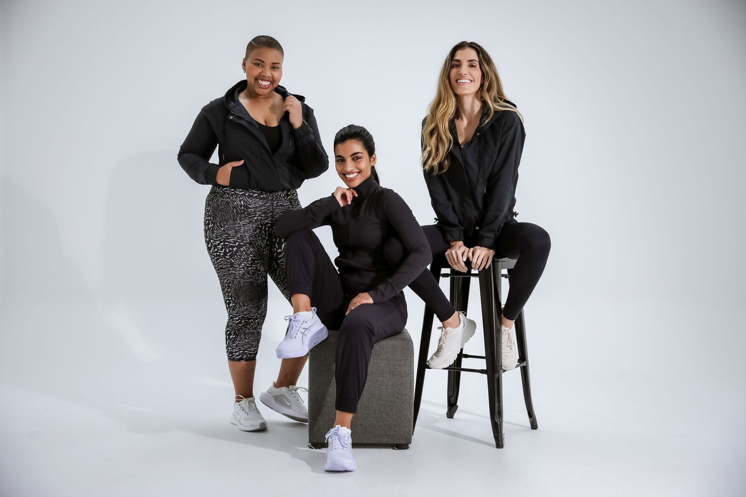 Three women in black athletic wear pose together in a bright white studio setting.