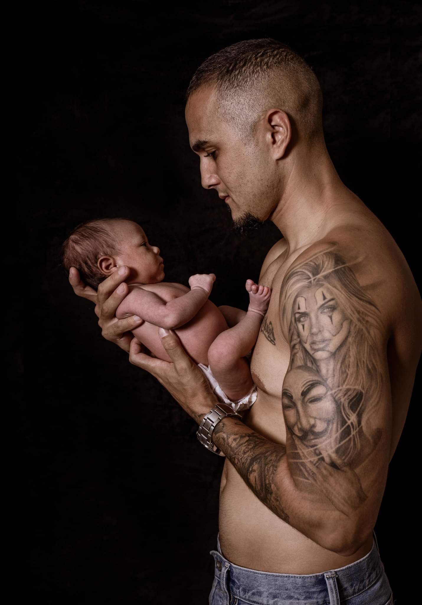 Tattooed shirtless father gently holds newborn baby in dramatic dark studio portrait photography.