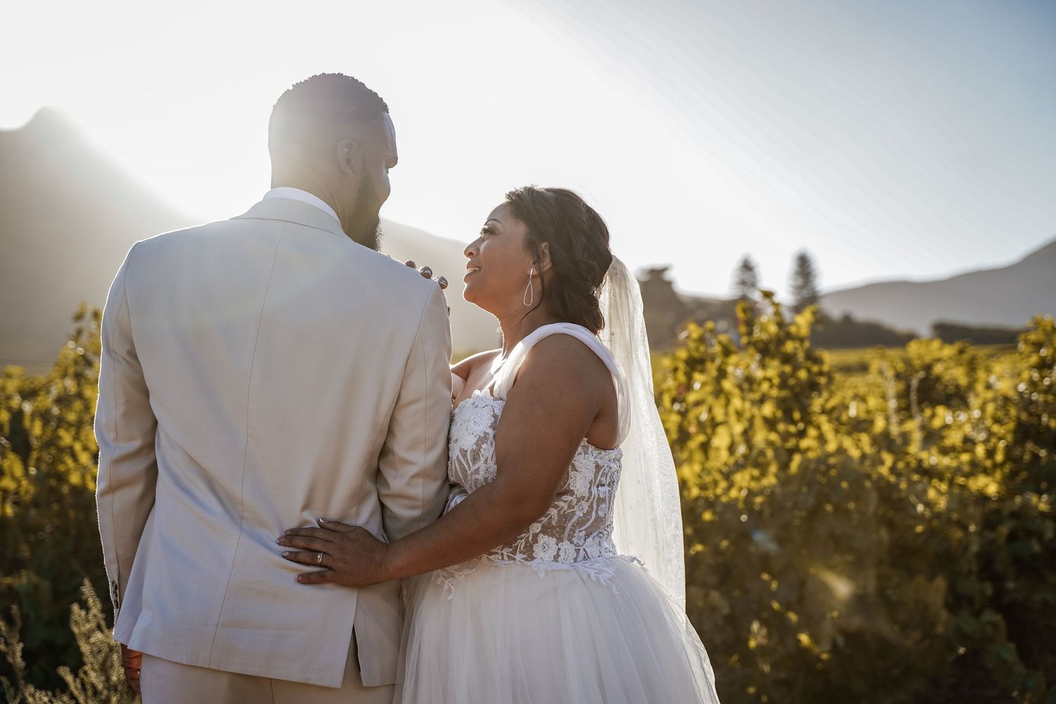 Bride and groom embrace while gazing at each other in a sunlit vineyard on their wedding day.