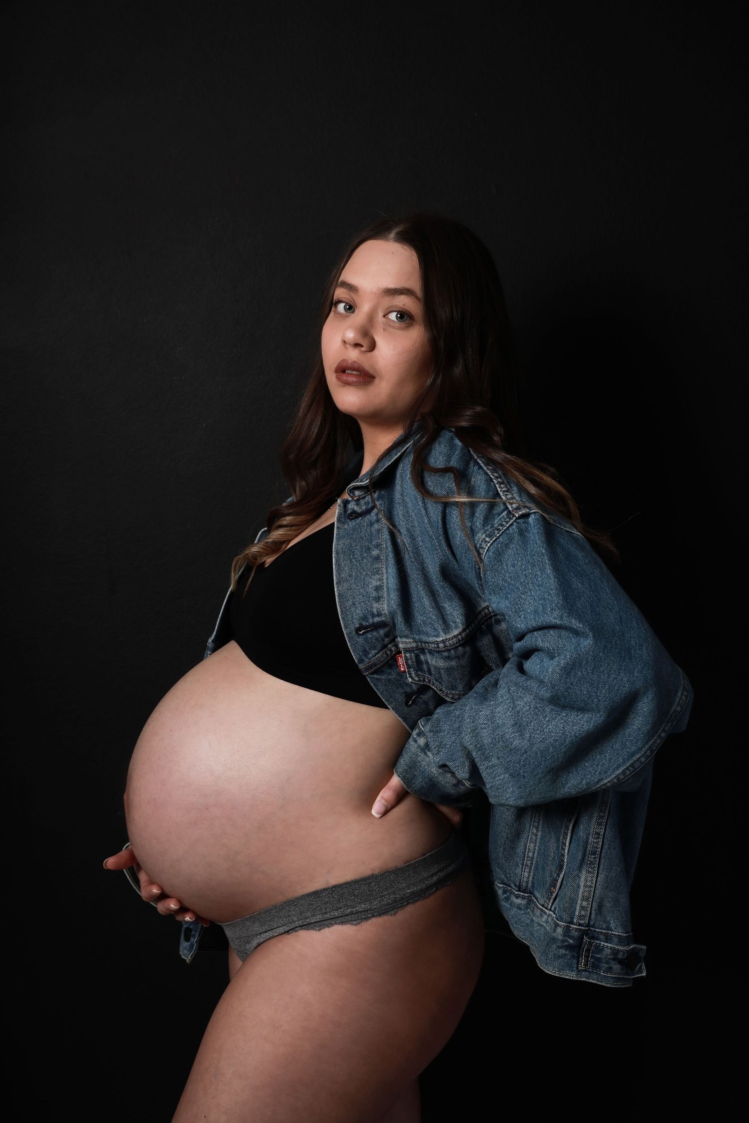 Pregnant woman in denim jacket and black crop top holds baby bump against dark background.
