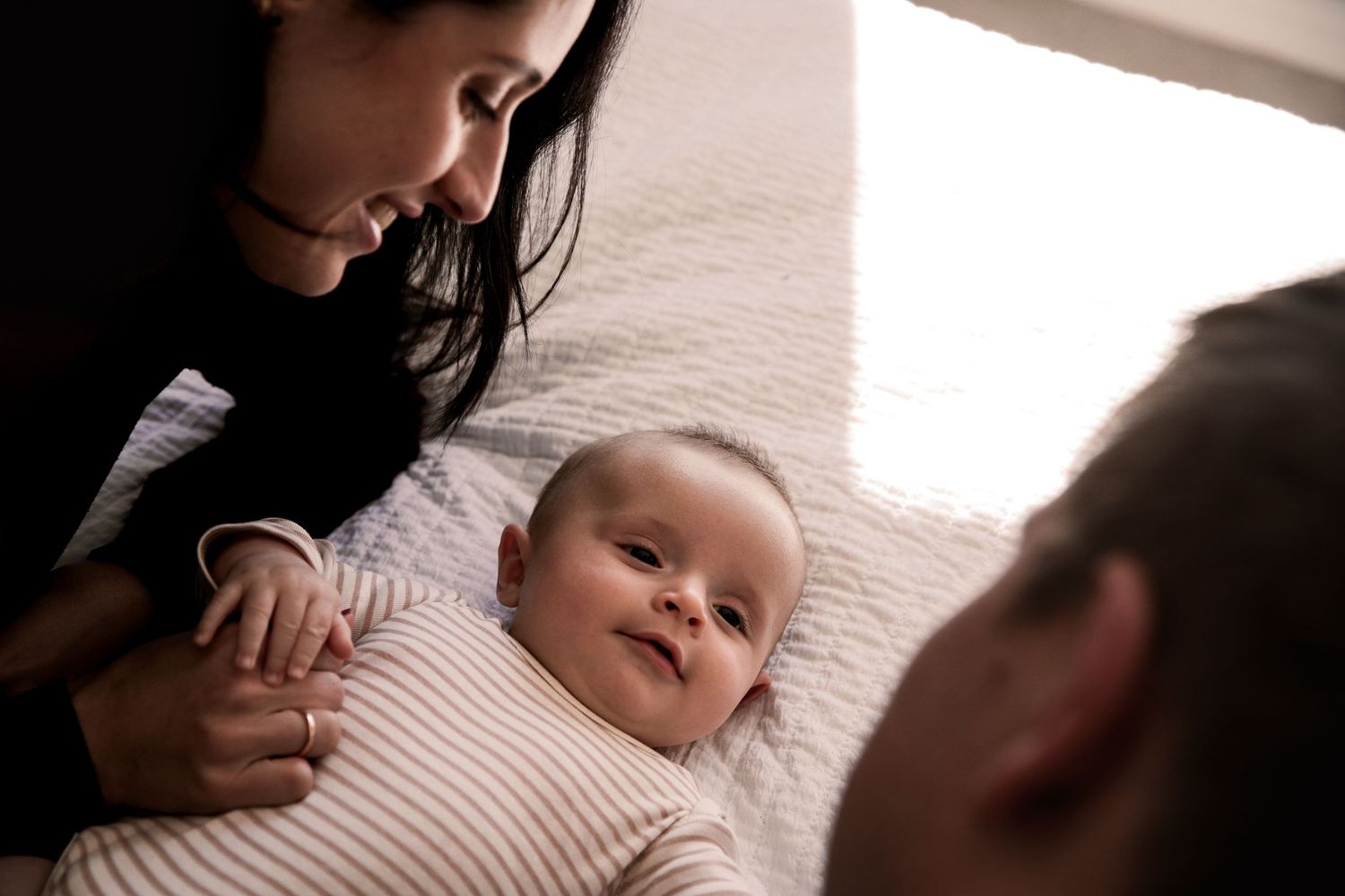 Smiling baby lying on a white blanket while parents lean in affectionately.