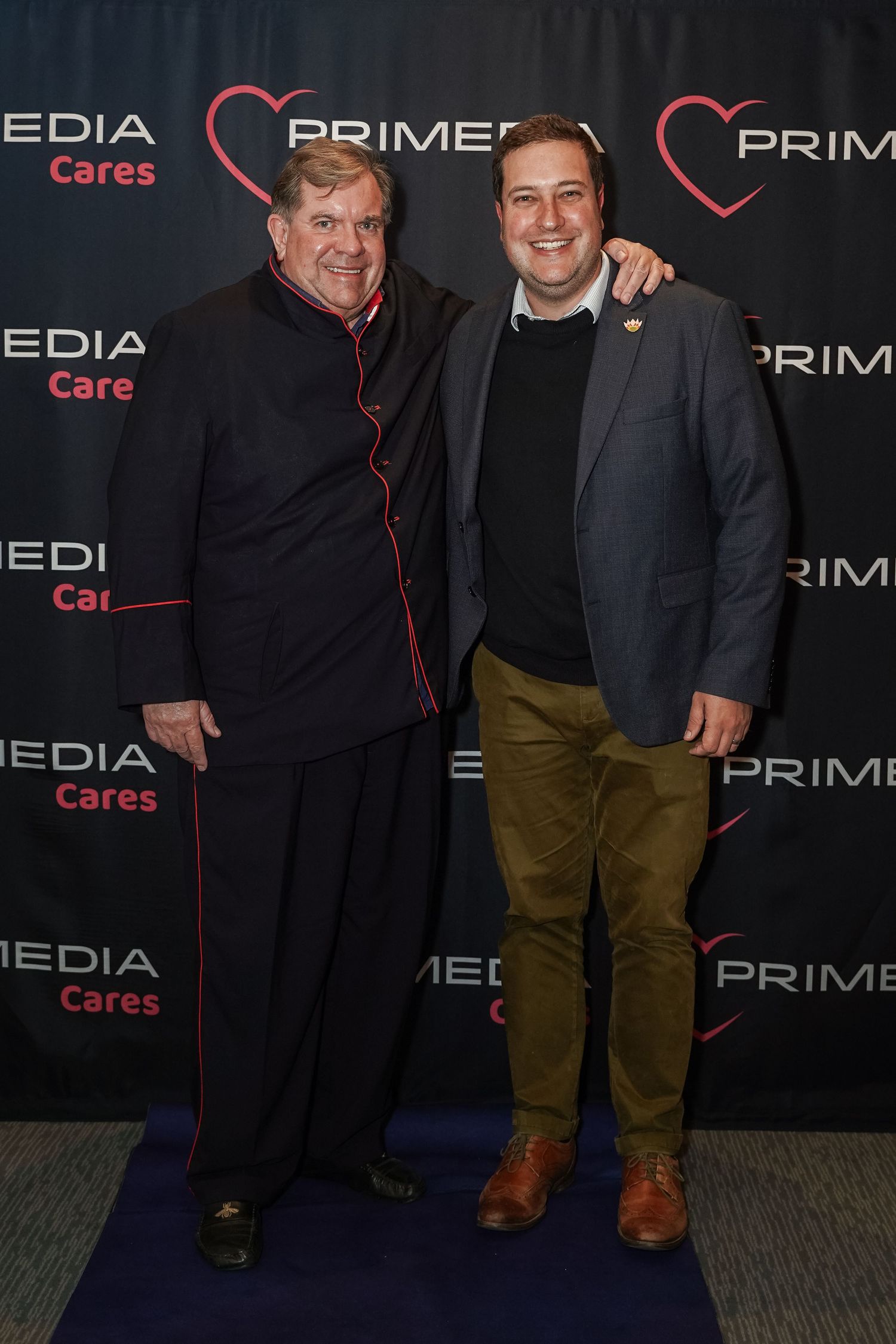 Two men posing together in front of a Primedia Cares event backdrop.
