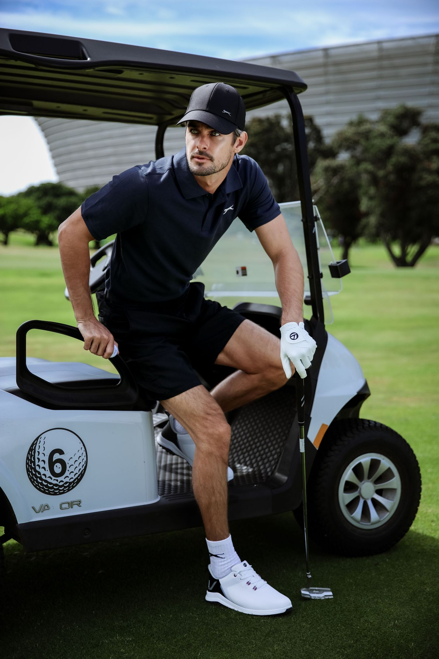 Male golfer in navy polo and shorts leans against white golf cart on sunny course.