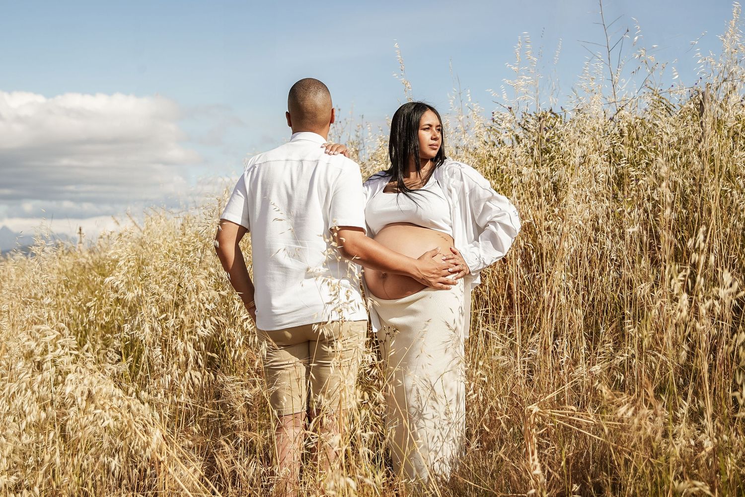 A pregnant woman in white skirt and her partner walk hand in hand through a golden wheat field under cloudy sky.