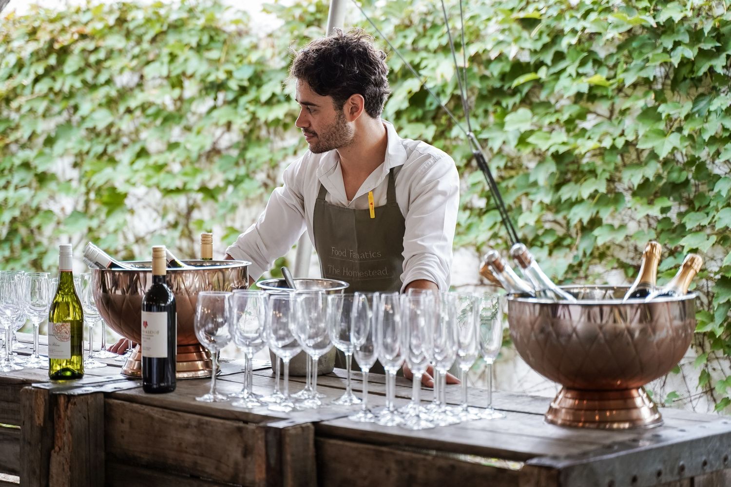 Bartender in apron arranging wine glasses and bottles at an outdoor bar with copper champagne buckets.