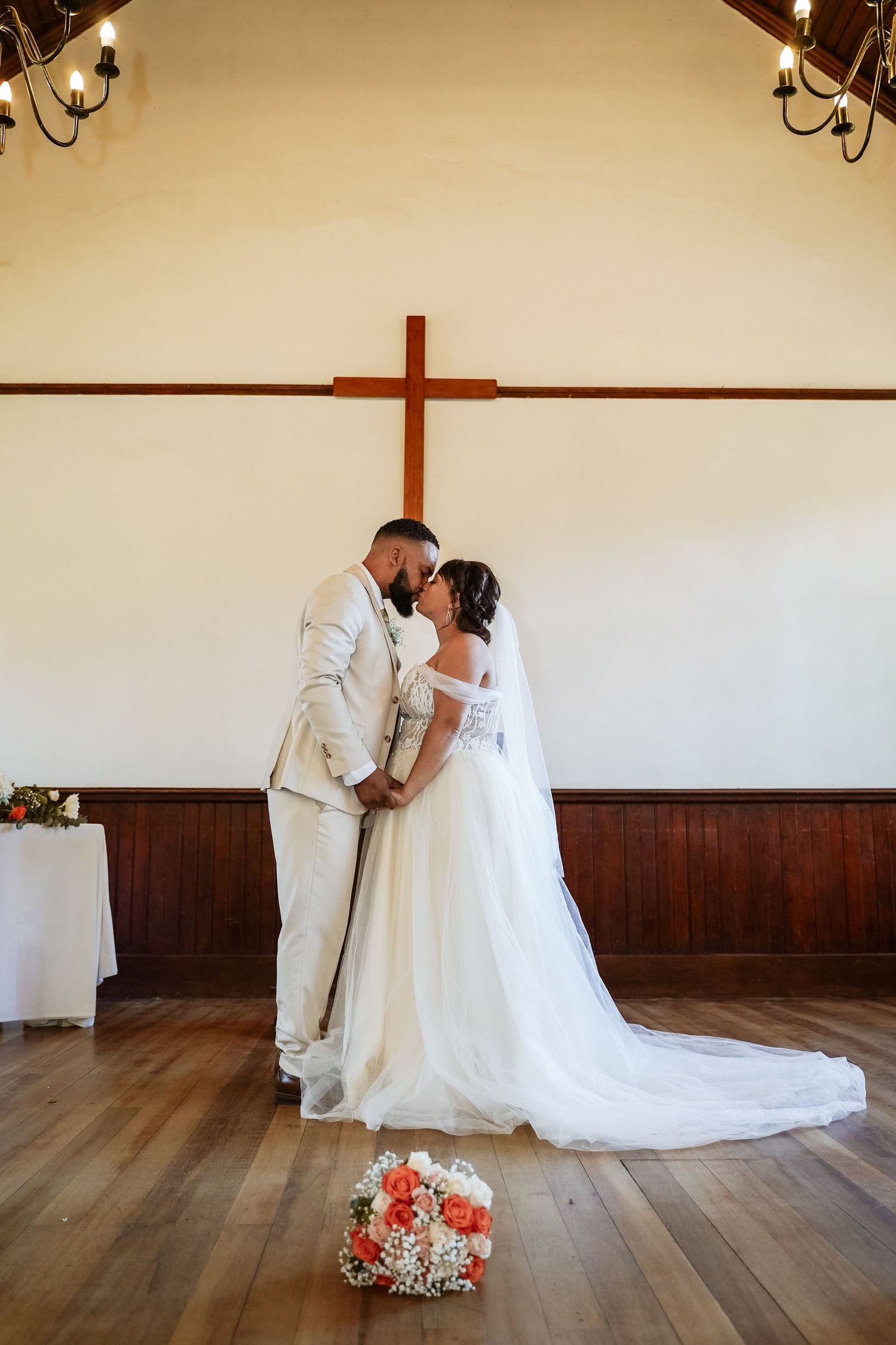 Newlyweds sharing first kiss in rustic chapel with wooden cross, bouquet on hardwood floor.