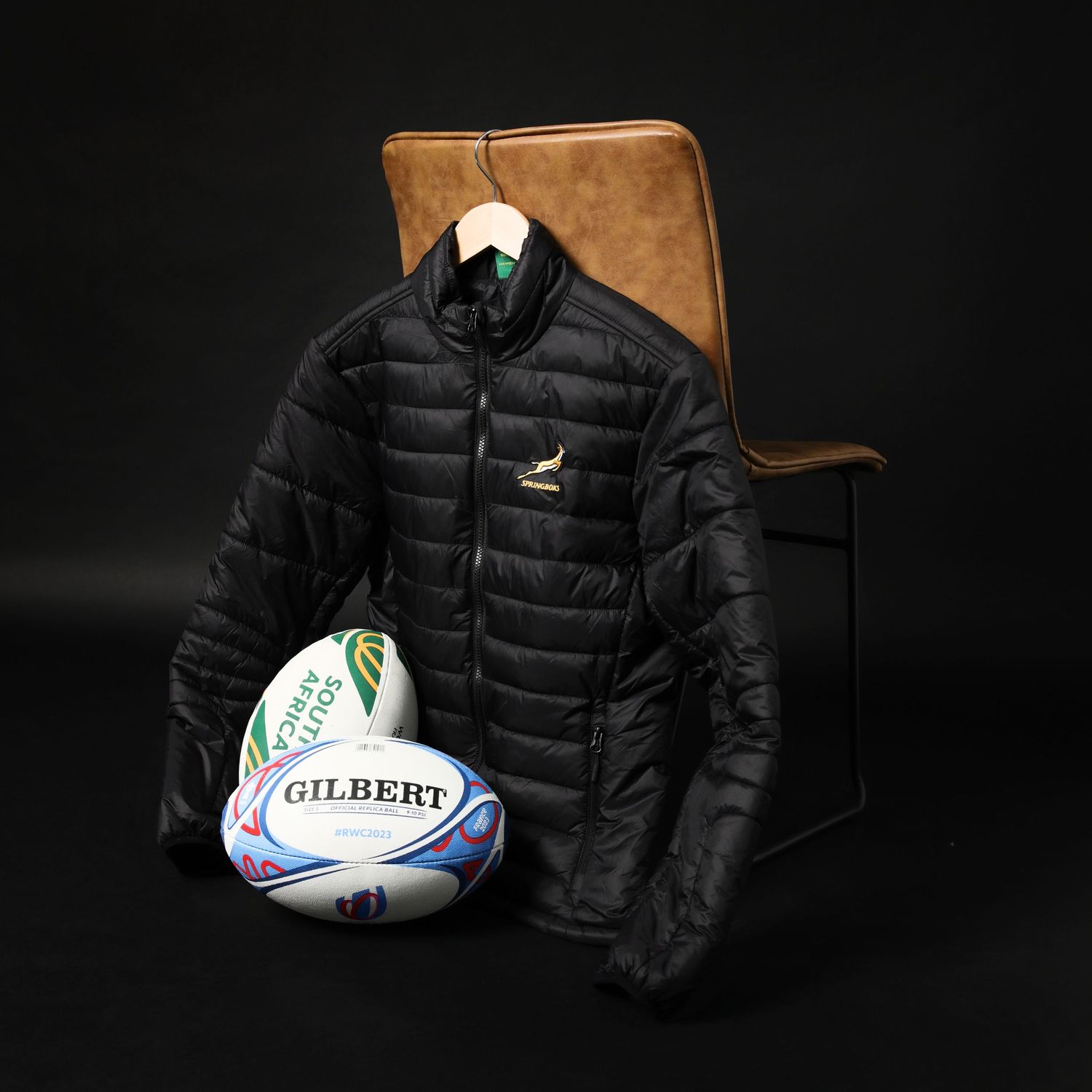 South Africa Springboks black puffer jacket displayed on chair with Gilbert rugby ball on dark background.