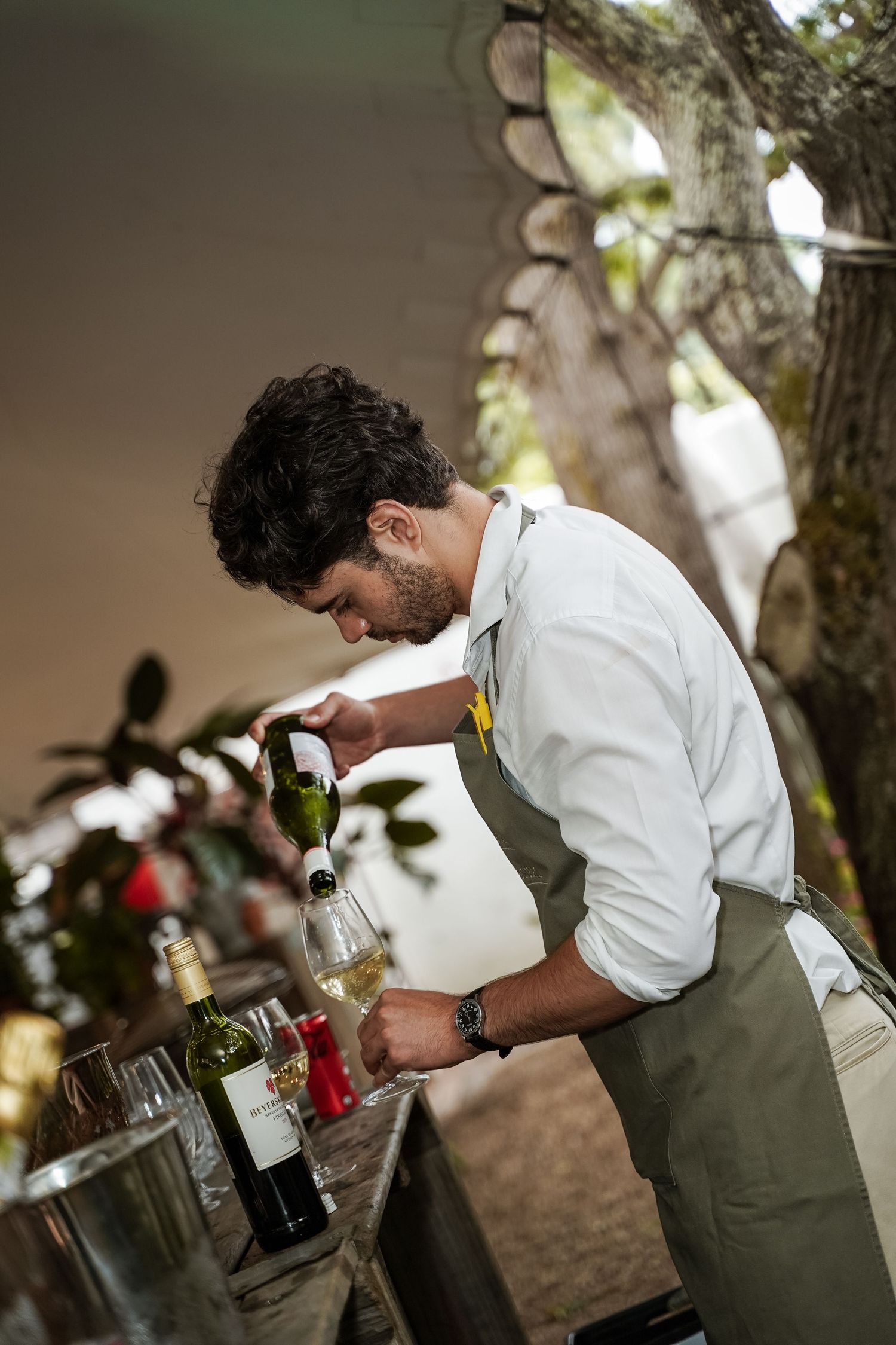 Male bartender in apron pouring white wine at an outdoor event bar surrounded by greenery.