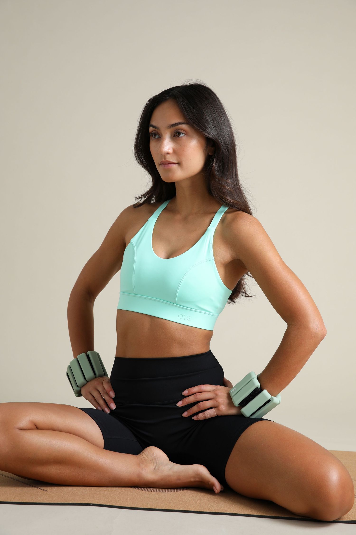 Athletic woman in mint sports bra and black shorts sits on mat holding green weights.