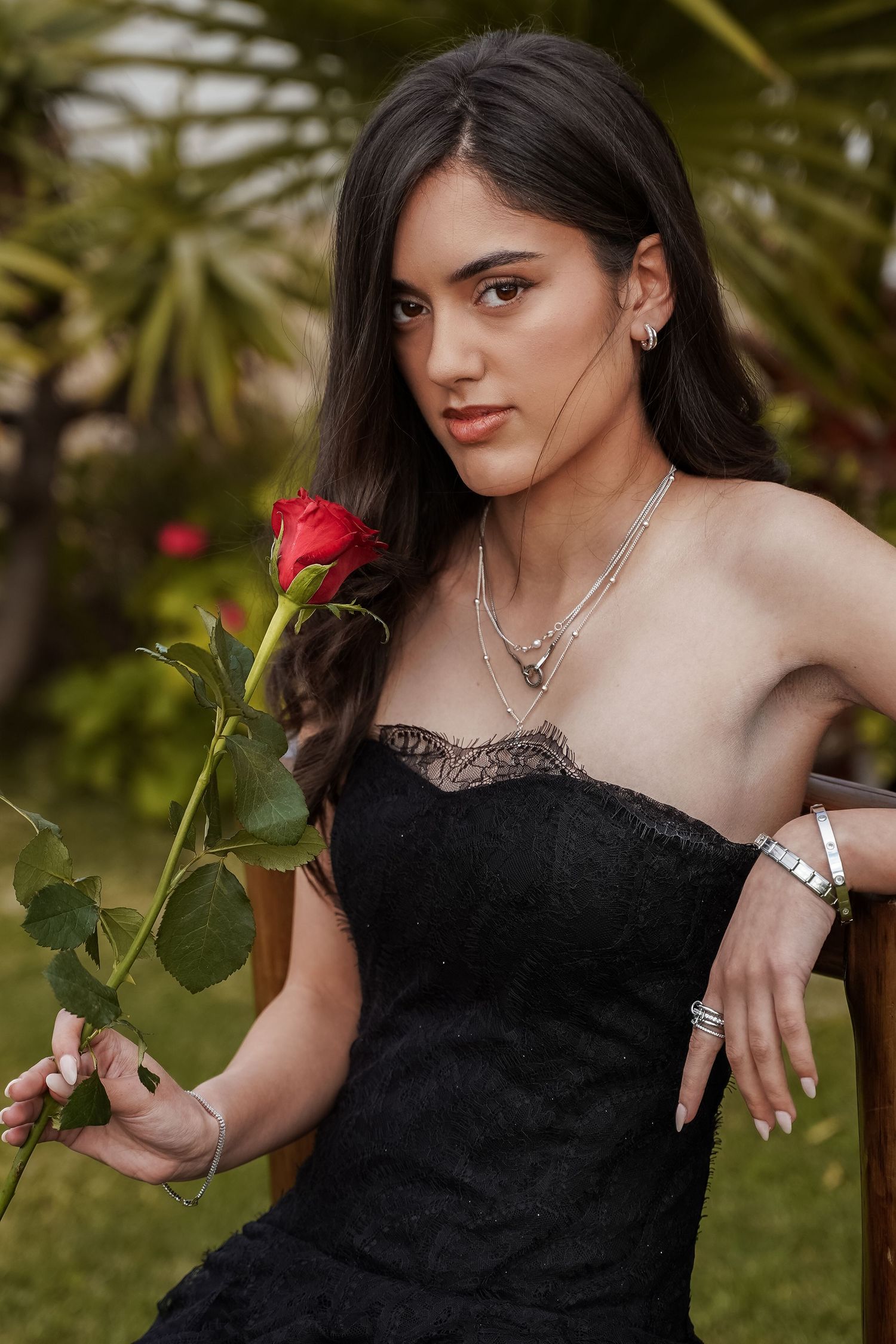 Young woman in black strapless lace dress holding a red rose while seated outdoors in garden setting.
