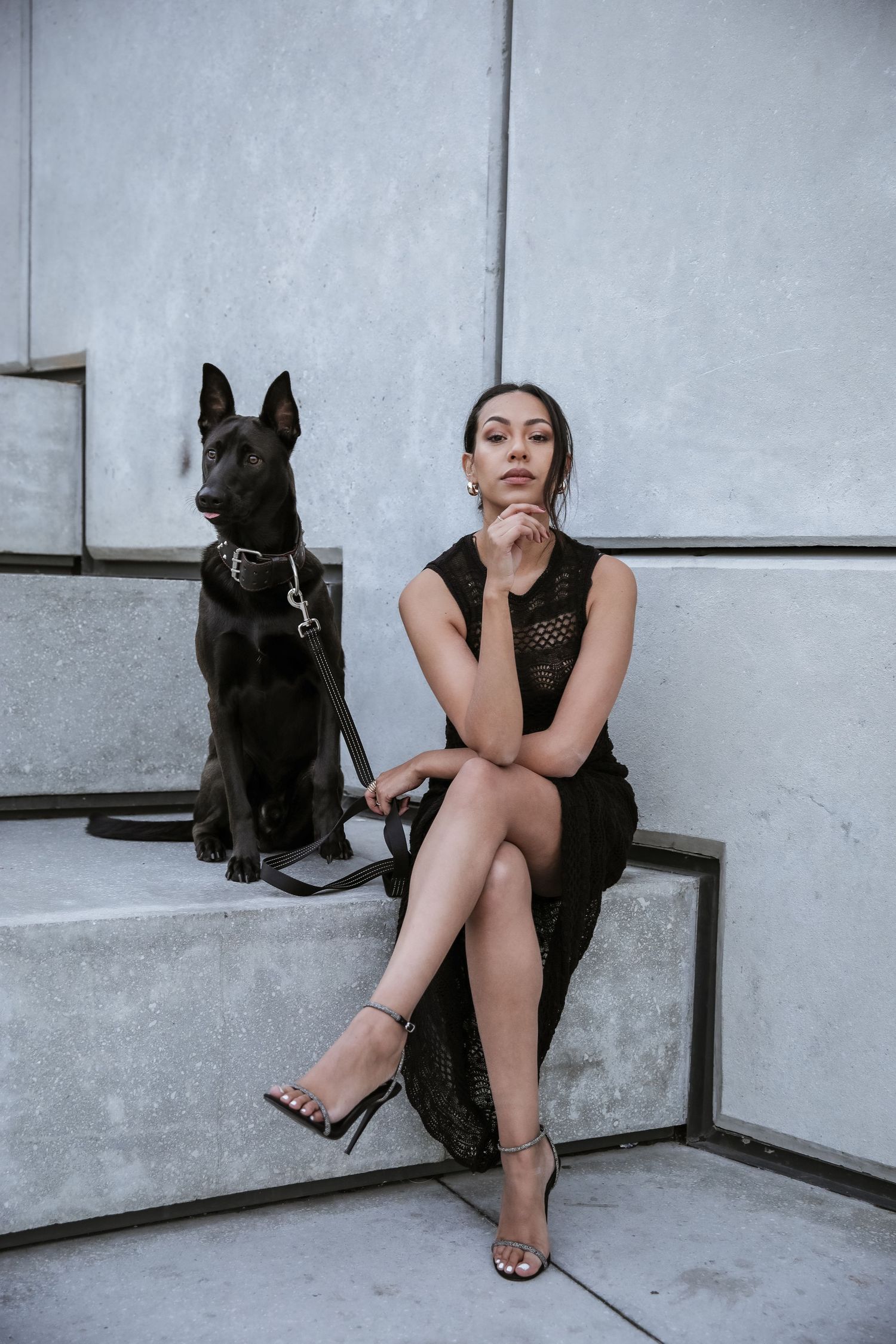 Woman in black lace dress sitting on steps with black German Shepherd dog on leash.
