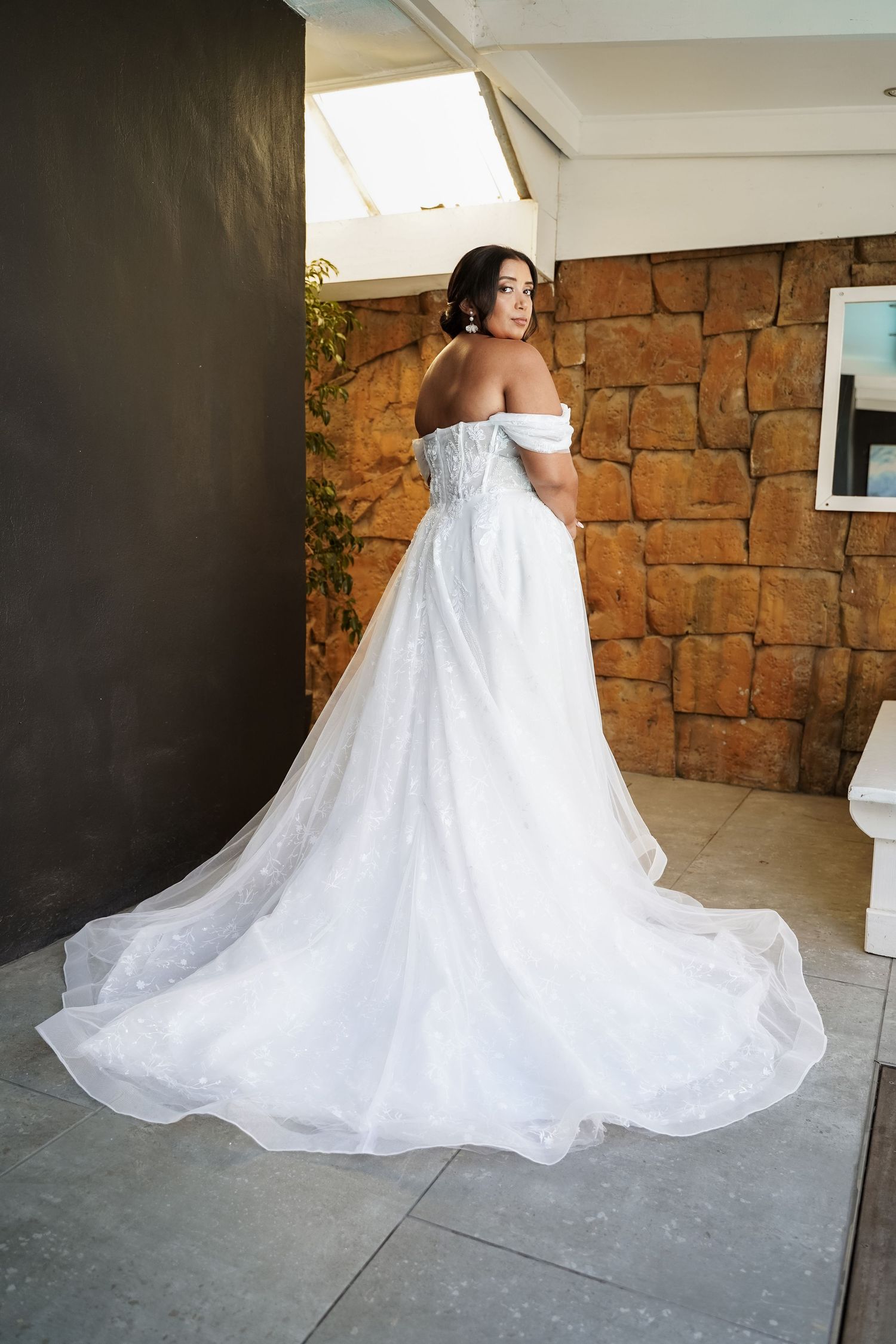Bride in off-shoulder white ball gown with long train, posing against rustic stone wall.