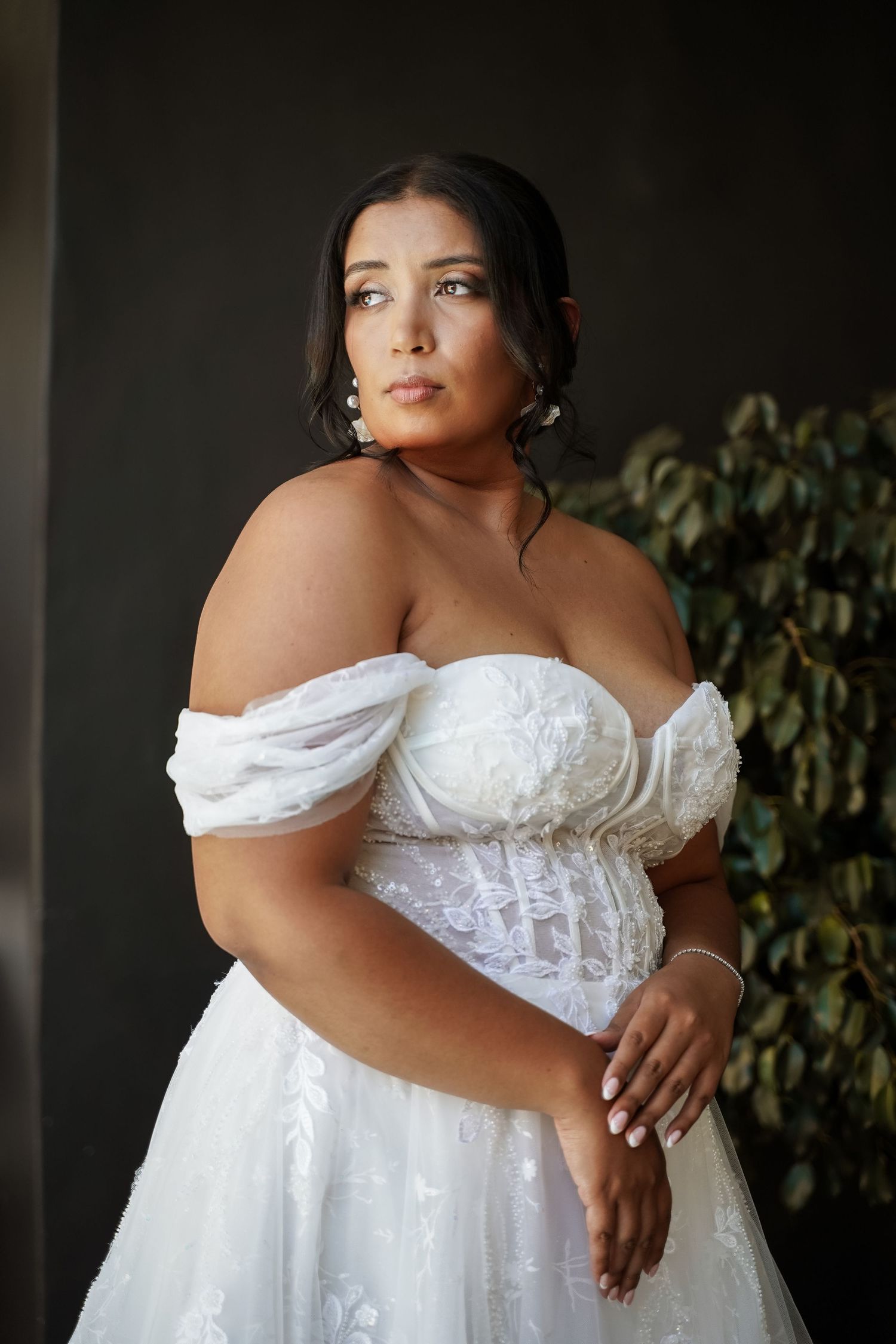 Beautiful plus-size bride in off-shoulder white lace wedding gown posing against dark background.