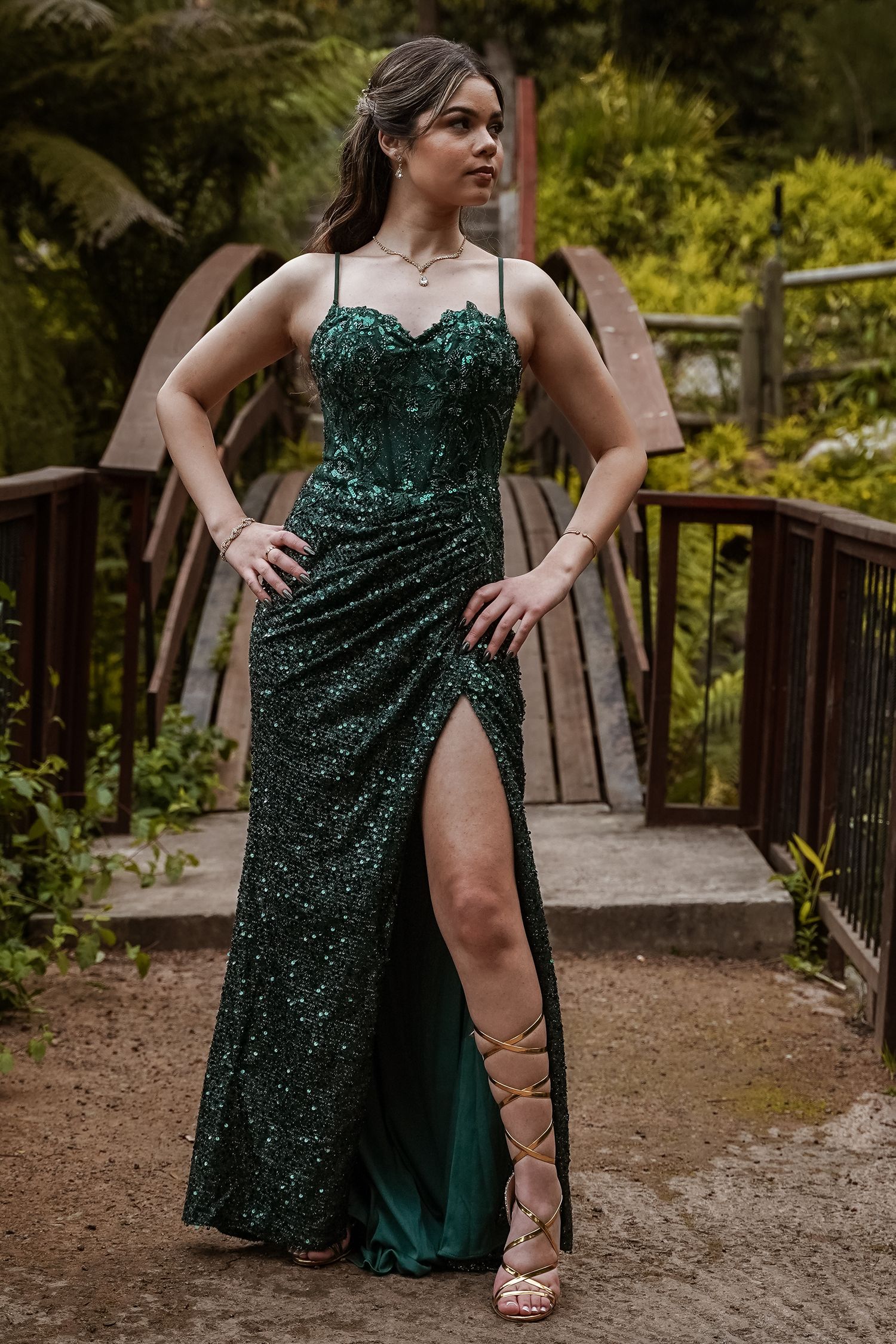 Young woman in emerald sequin gown with thigh slit and lace-up heels poses on rustic wooden bridge.