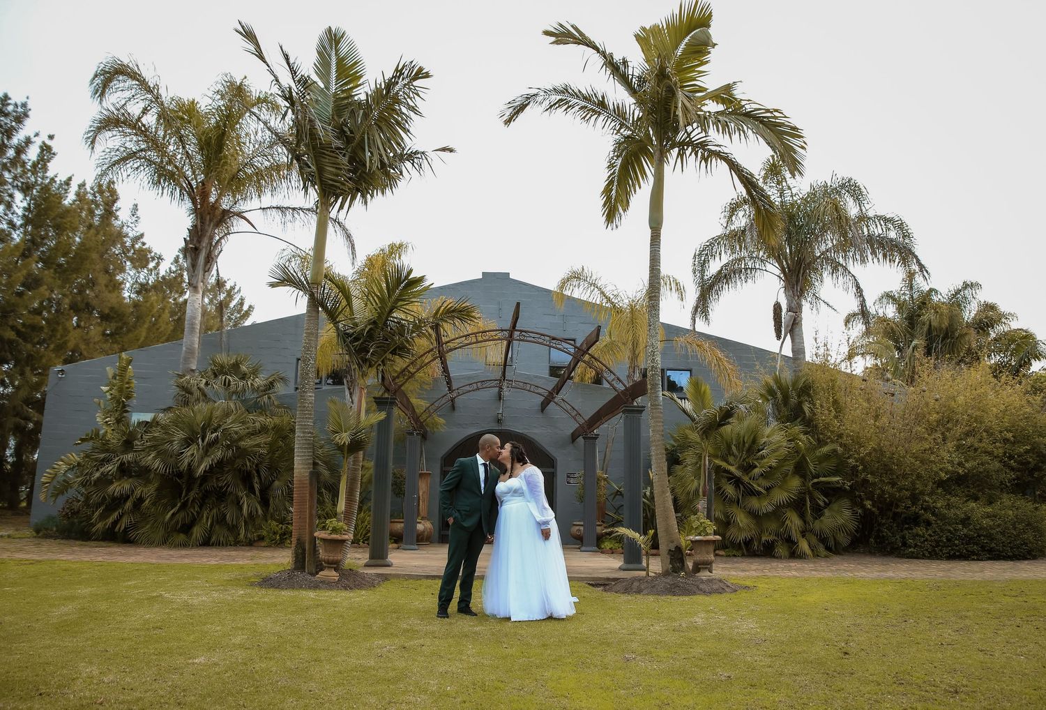 Newlywed couple kissing on green lawn surrounded by tall palm trees outside elegant venue.