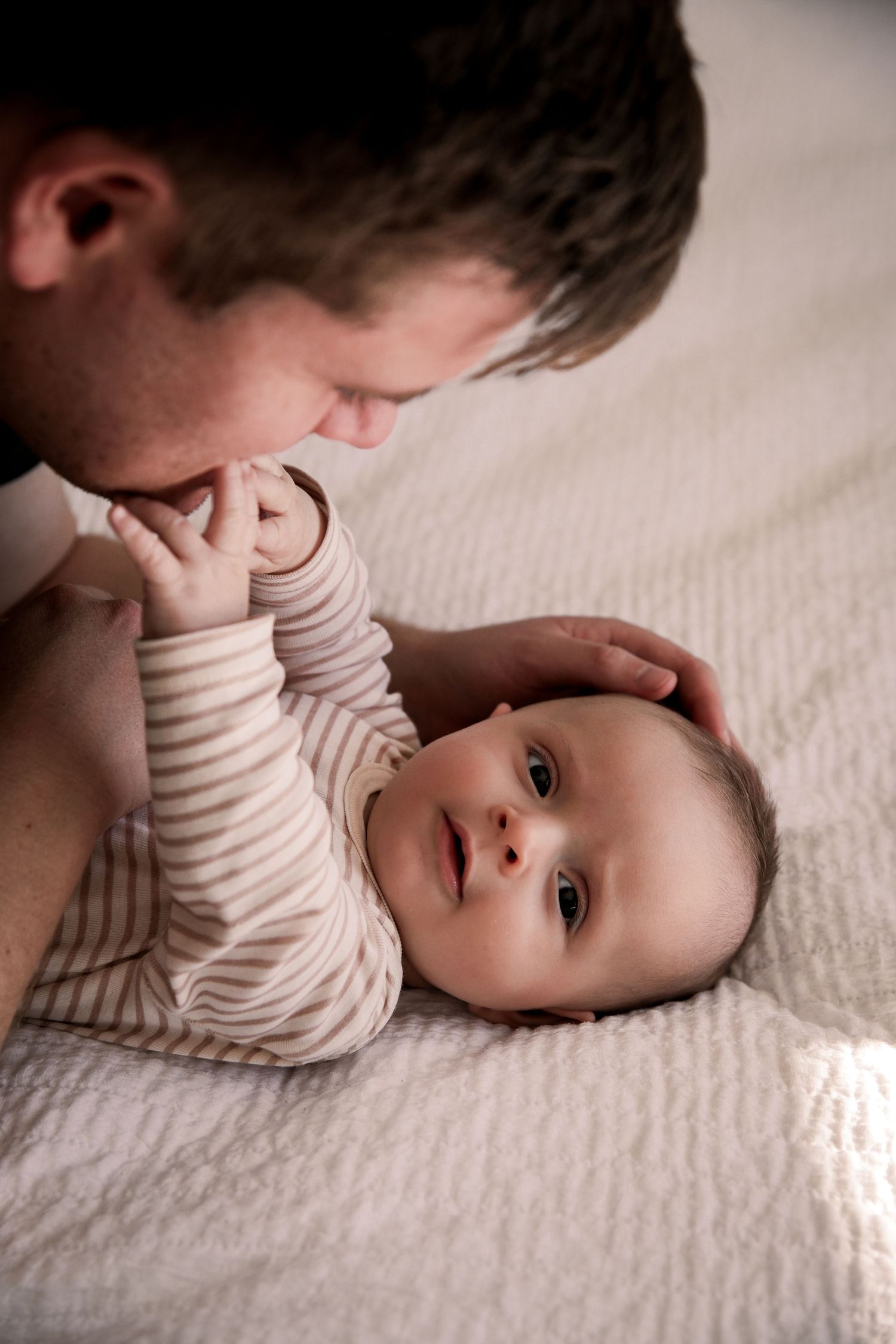 Father gently cradling his alert baby lying on a soft white quilted bed.