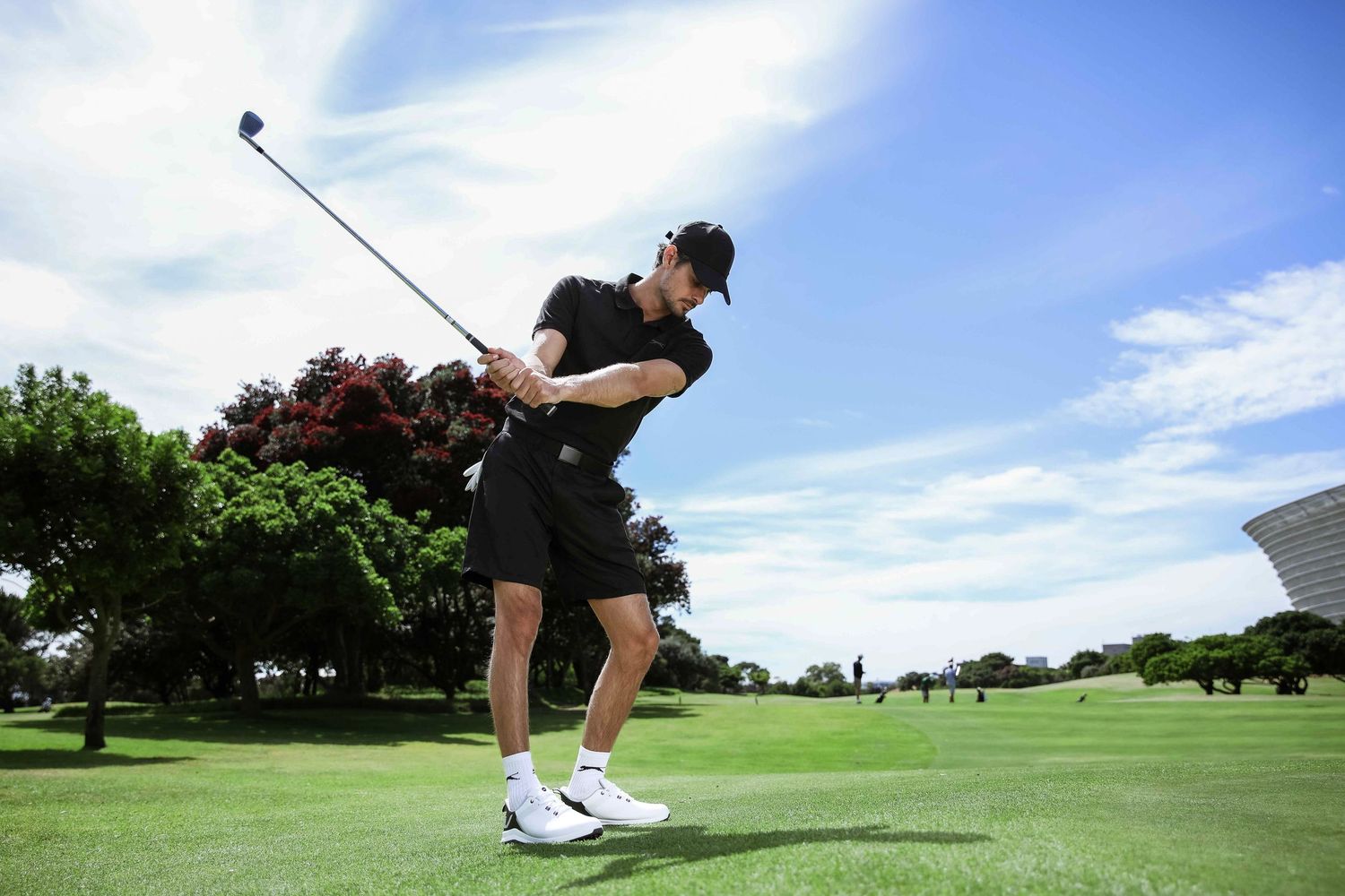 Male golfer in black outfit swings club on bright sunny golf course.