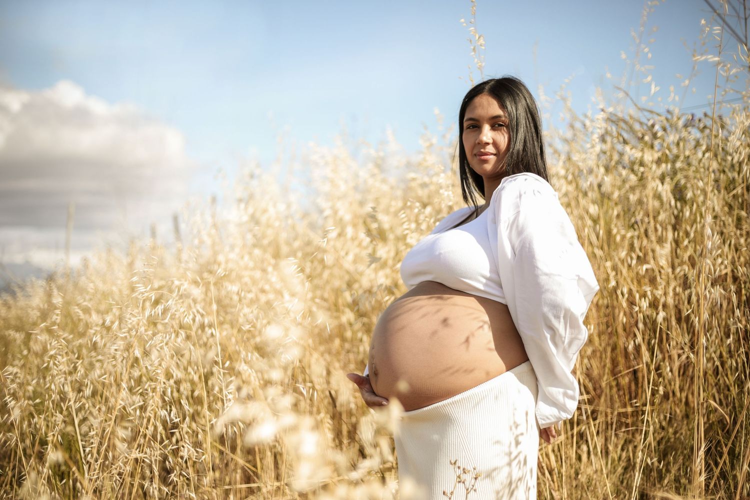A pregnant woman in white crop top and skirt cradles her bare belly while standing in a golden wheat field outdoors.