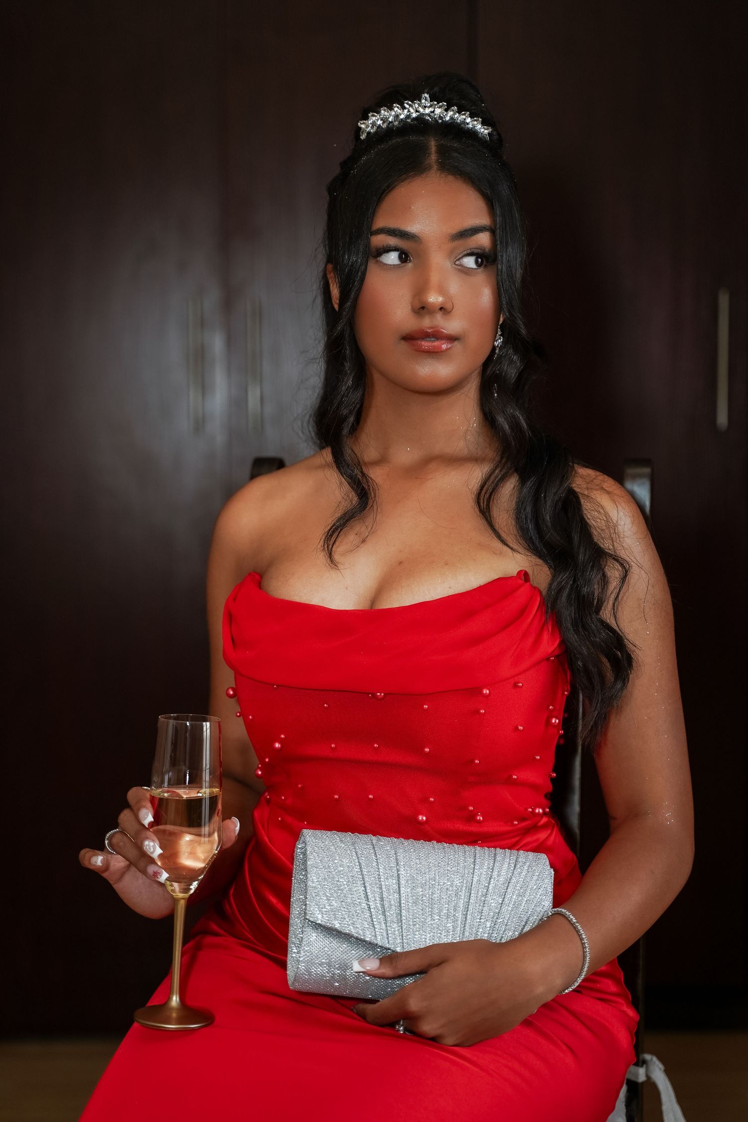 Young woman in red strapless gown with tiara holds champagne glass and silver clutch indoors.