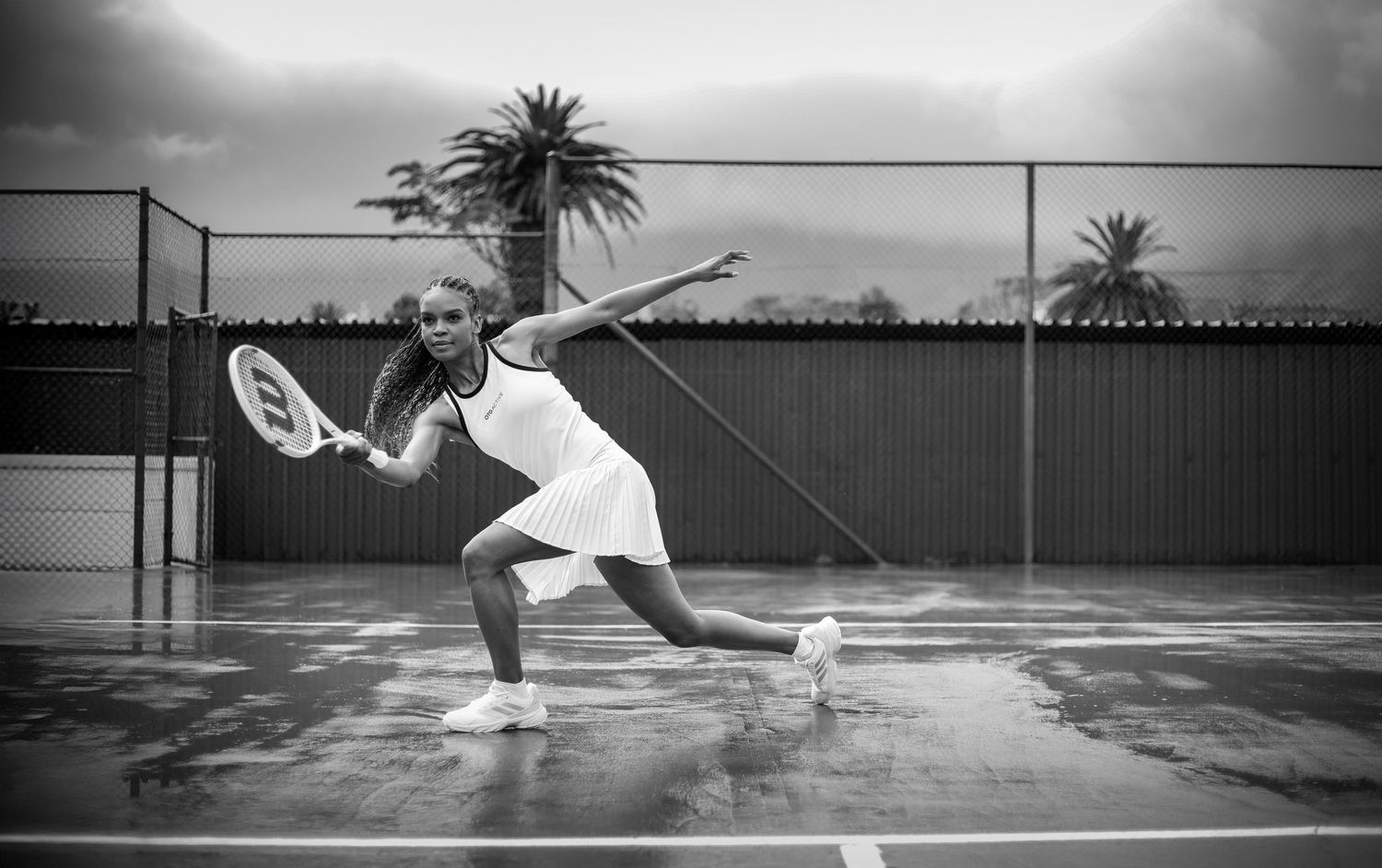Black and white photo of a woman playing tennis on a wet outdoor court in a white dress.