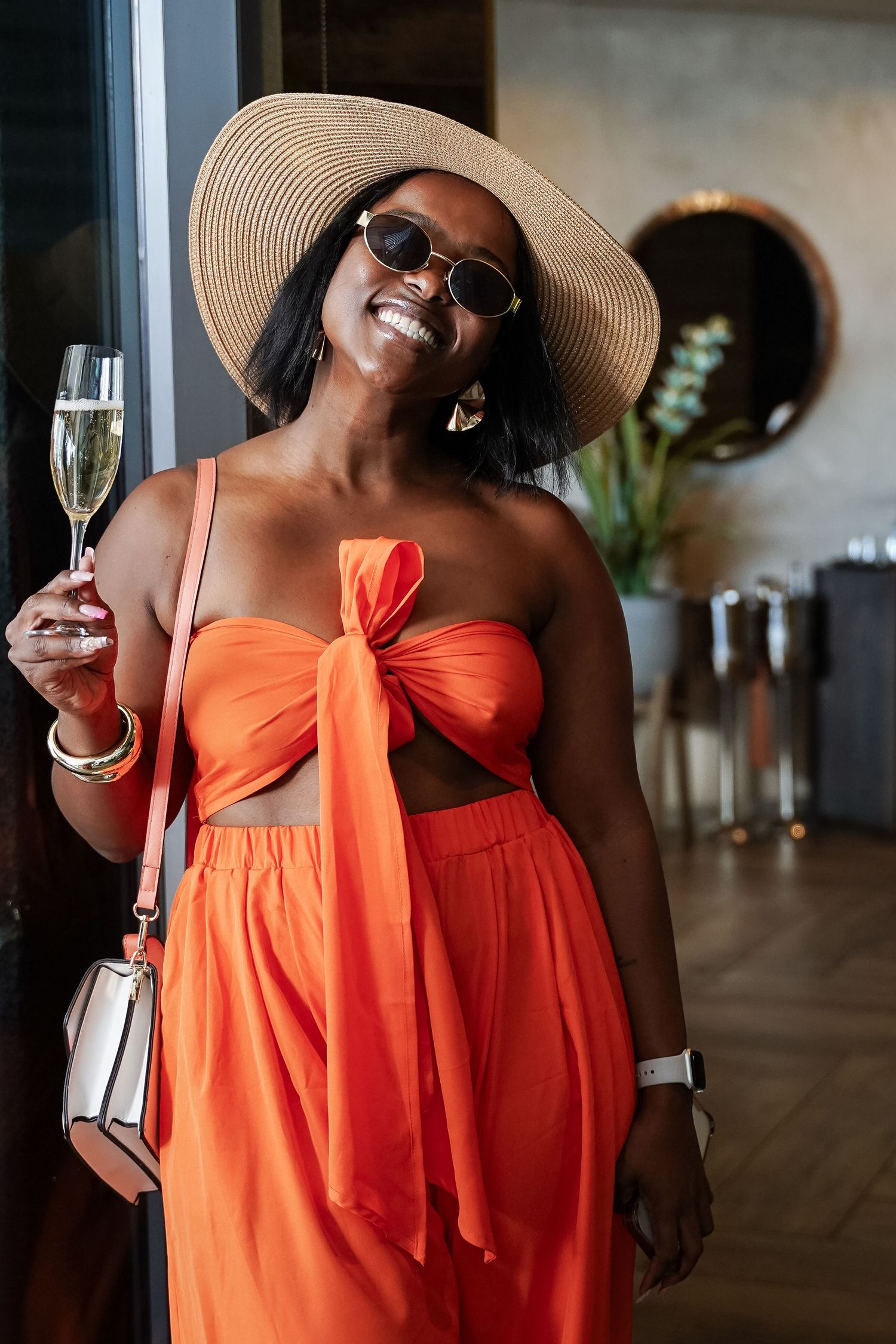 Stylish Black woman in orange two-piece outfit holding champagne glass wearing sun hat.