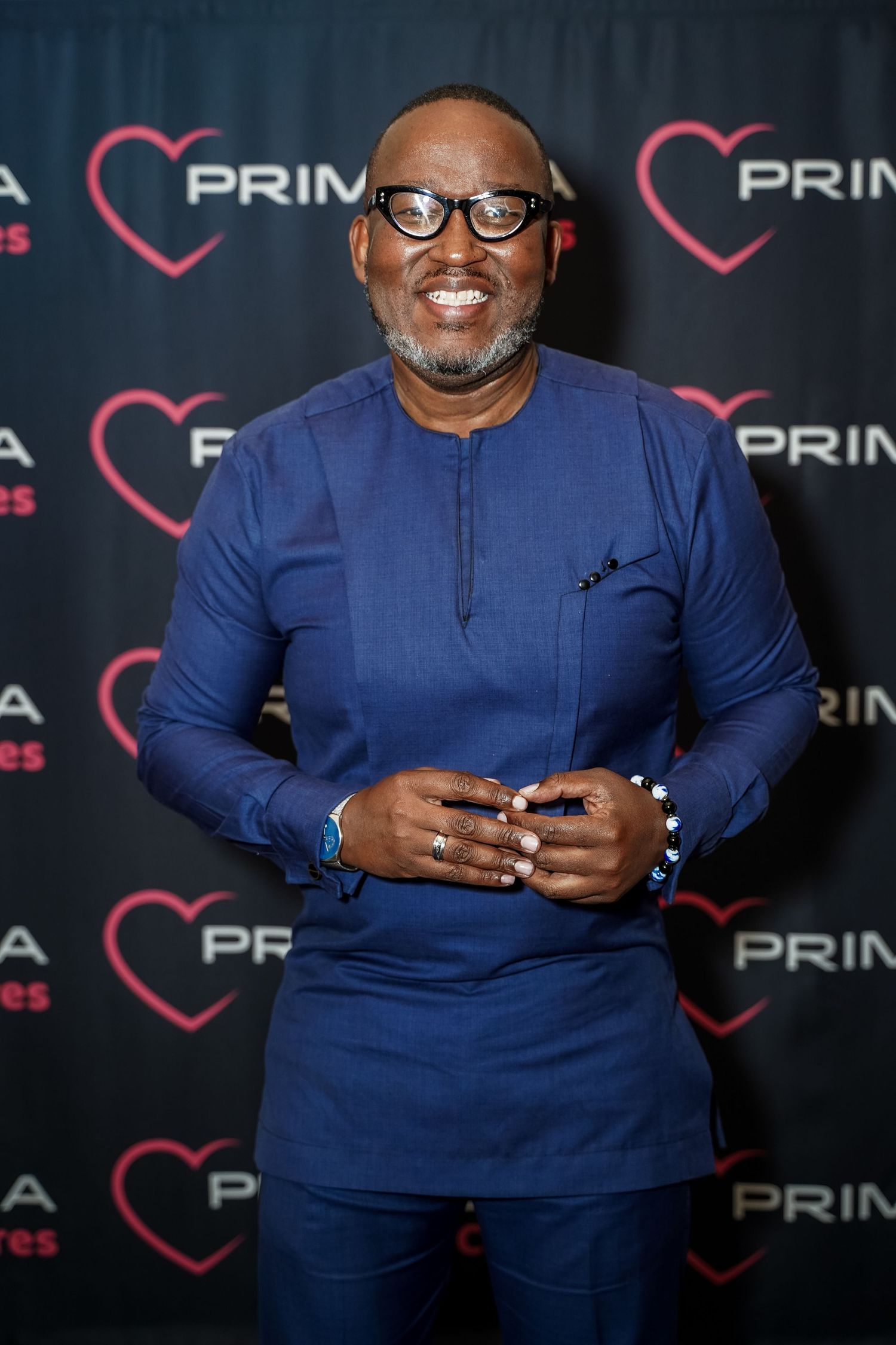 Elegantly dressed man in blue outfit smiling at Primedia Cares branded backdrop.