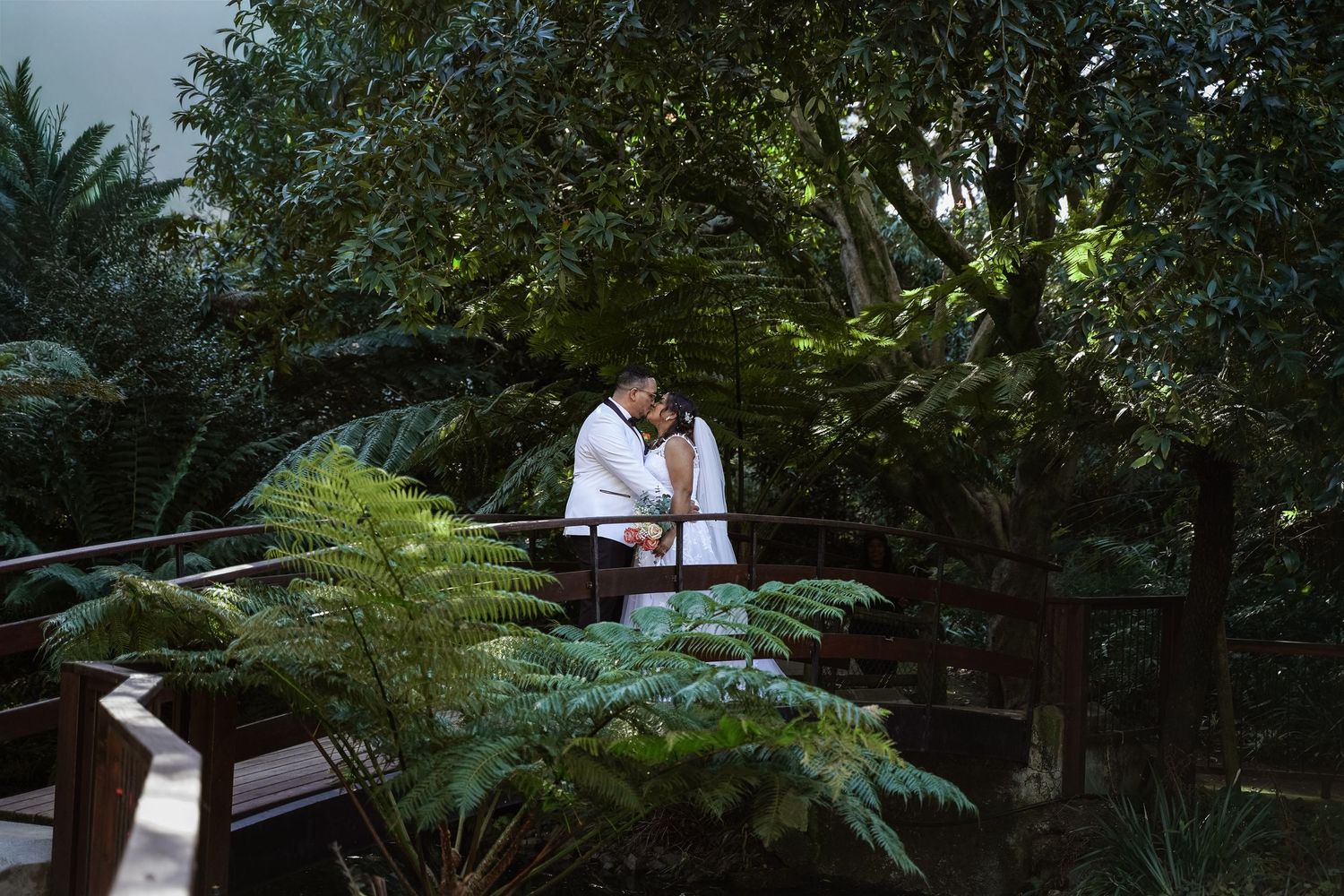 Couple sharing a romantic embrace on a wooden bridge surrounded by lush tropical greenery and ferns.