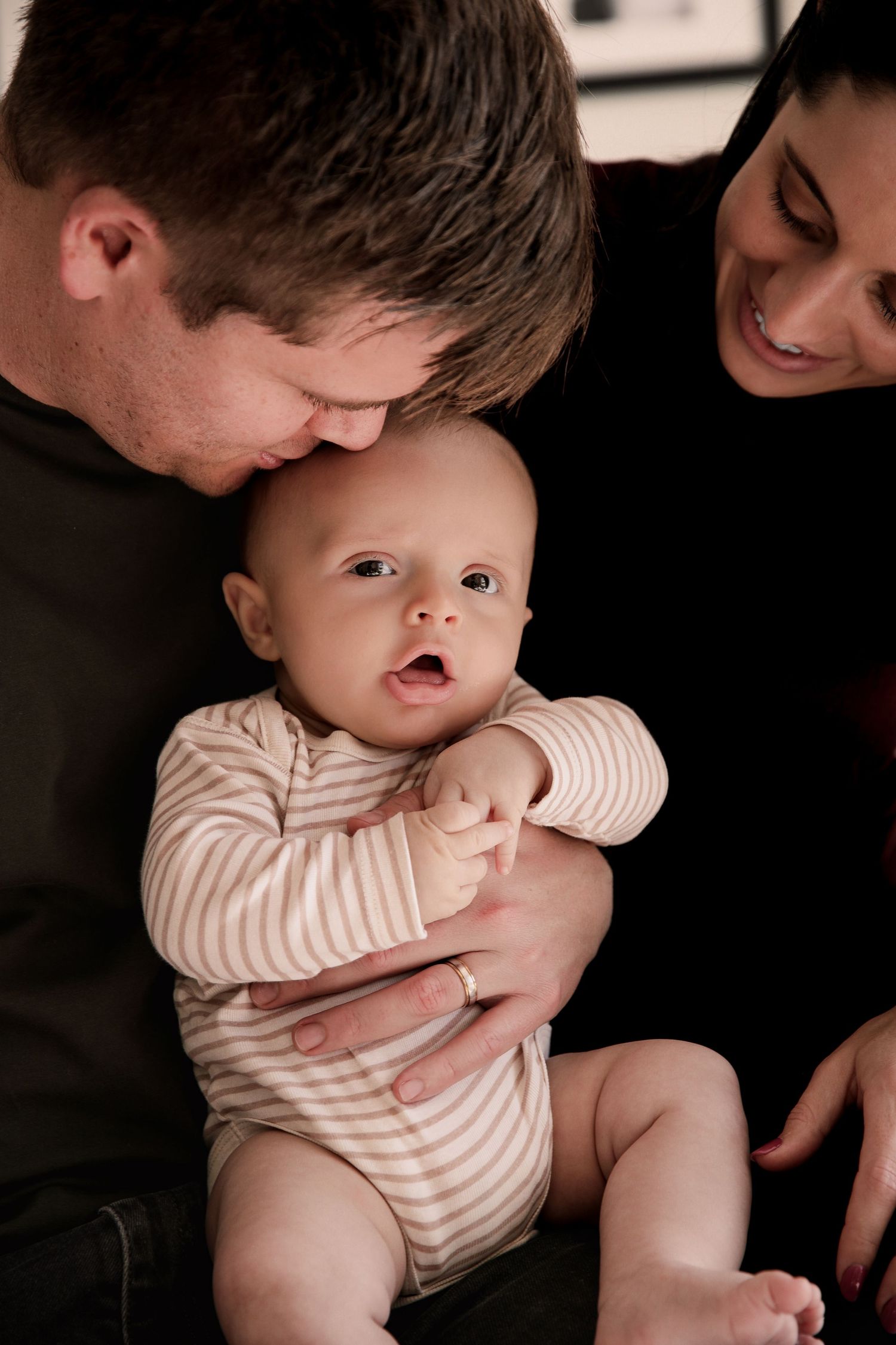 Loving parents in black outfits holding and smiling at their wide-eyed baby.