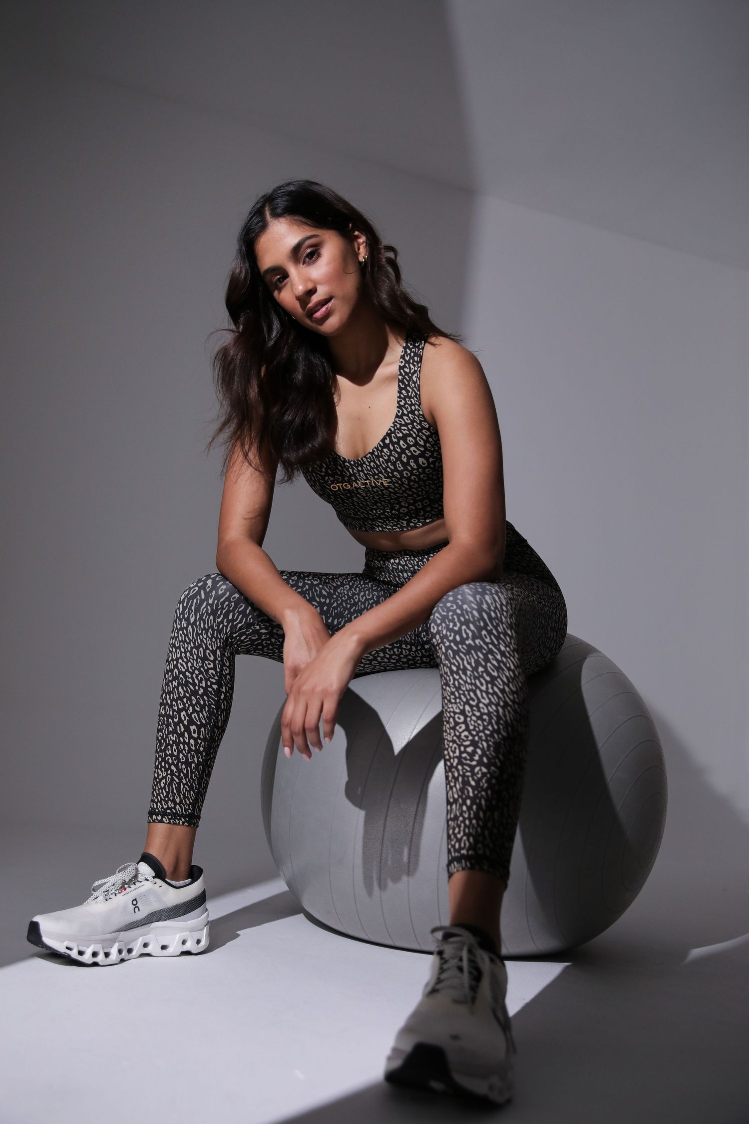 Woman in patterned grey activewear set sitting on a round prop wearing On Cloud sneakers.