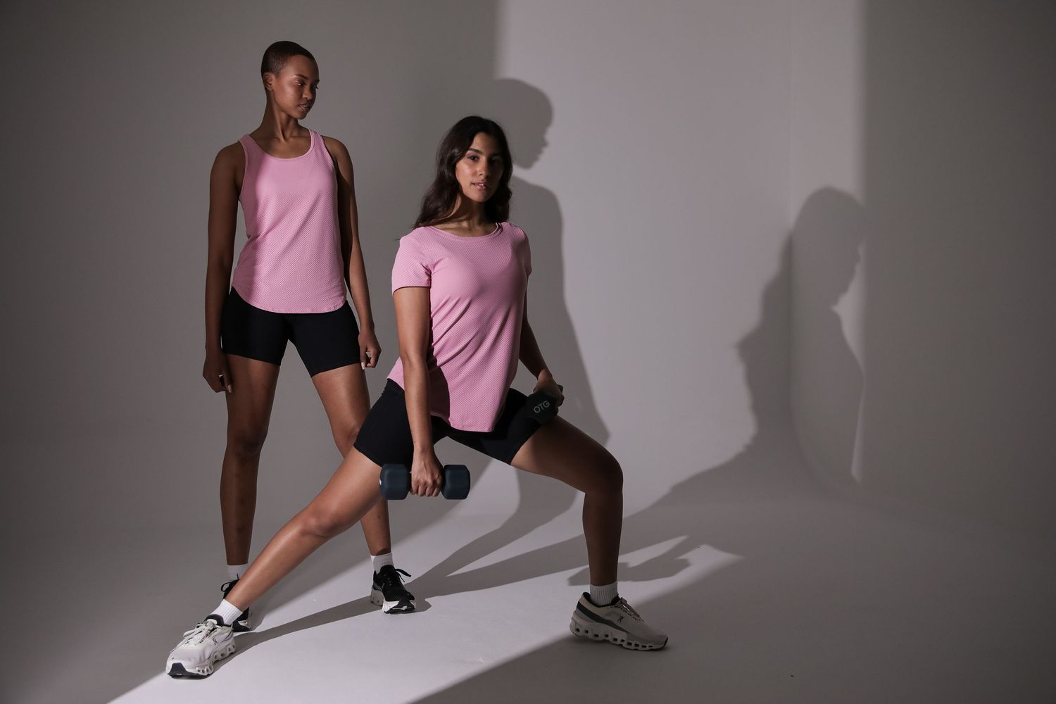 Two women in pink activewear and black shorts exercising with dumbbells in a studio.