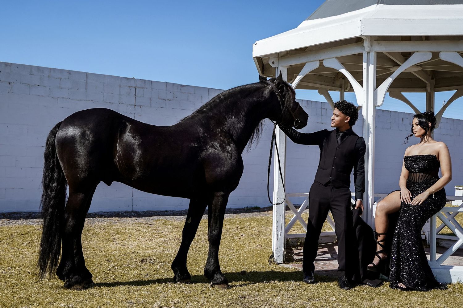 Couple in formal black attire posing with a black horse near a gazebo.