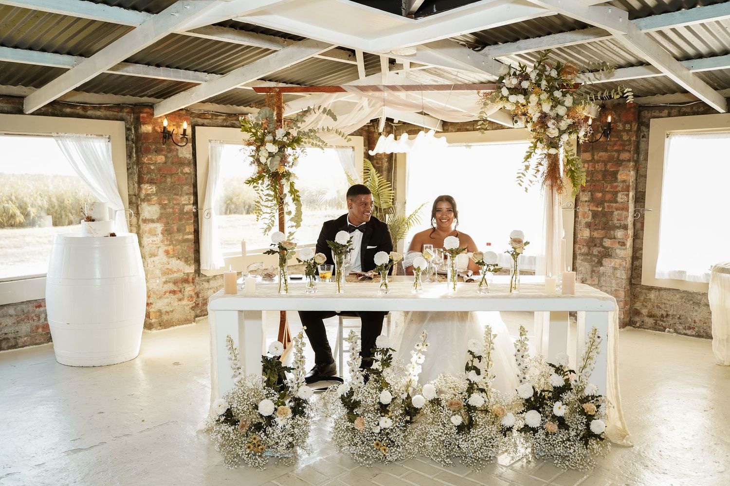 Wedding couple seated at elegant white sweetheart table adorned with white floral arrangements in rustic venue.
