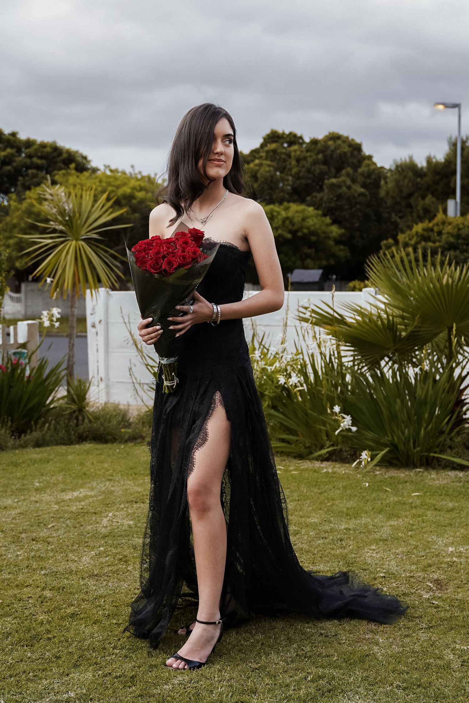 Young woman in black strapless tulle gown holding large bouquet of red roses standing in garden.
