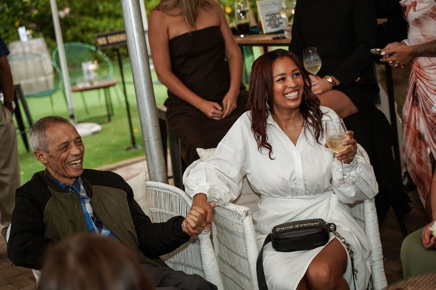 Joyful woman in white dress laughing and holding hands with an elderly man at an outdoor celebration.