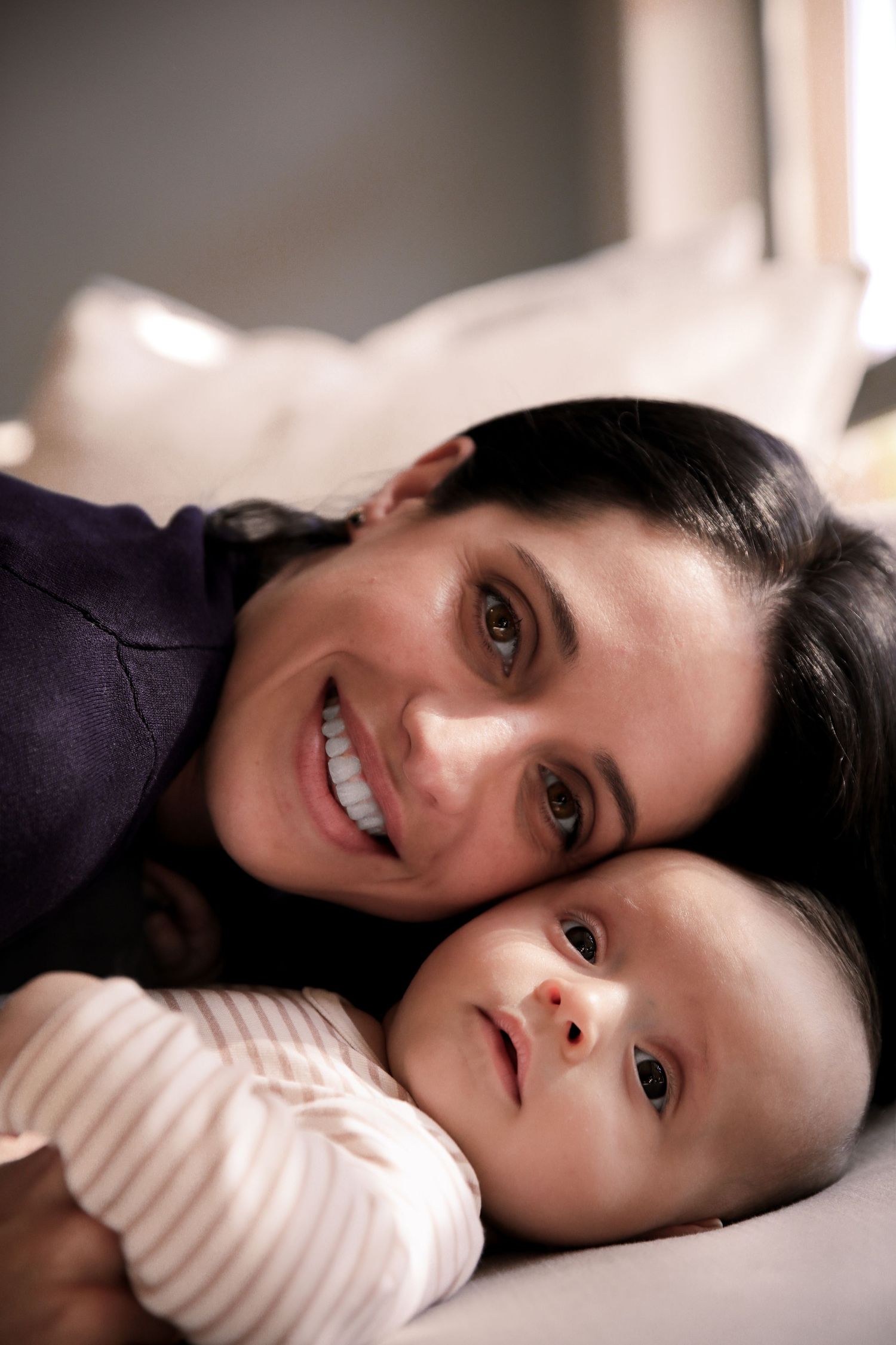 Smiling dark-haired mother lying cheek-to-cheek with her infant baby on a bed.