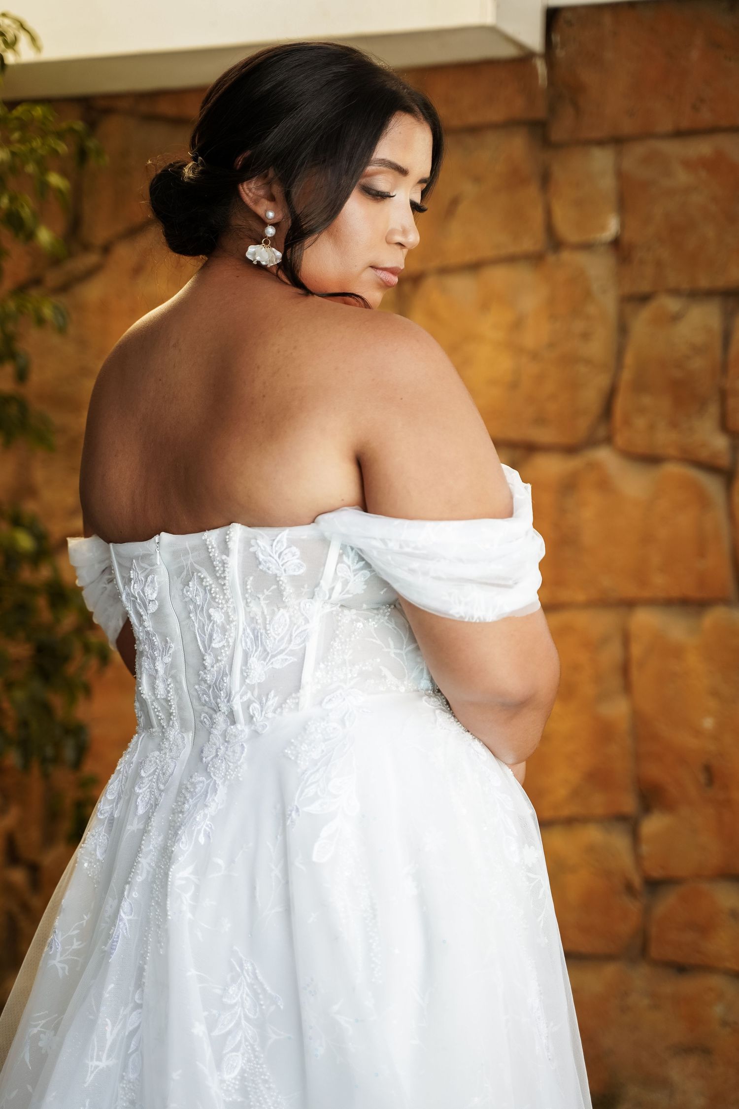 Bride in off-shoulder white lace wedding gown poses gracefully, showcasing elegant back detail and pearl earrings.