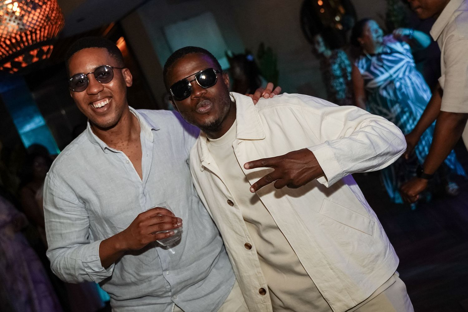Two smiling men posing together at a party, both wearing white outfits and sunglasses.