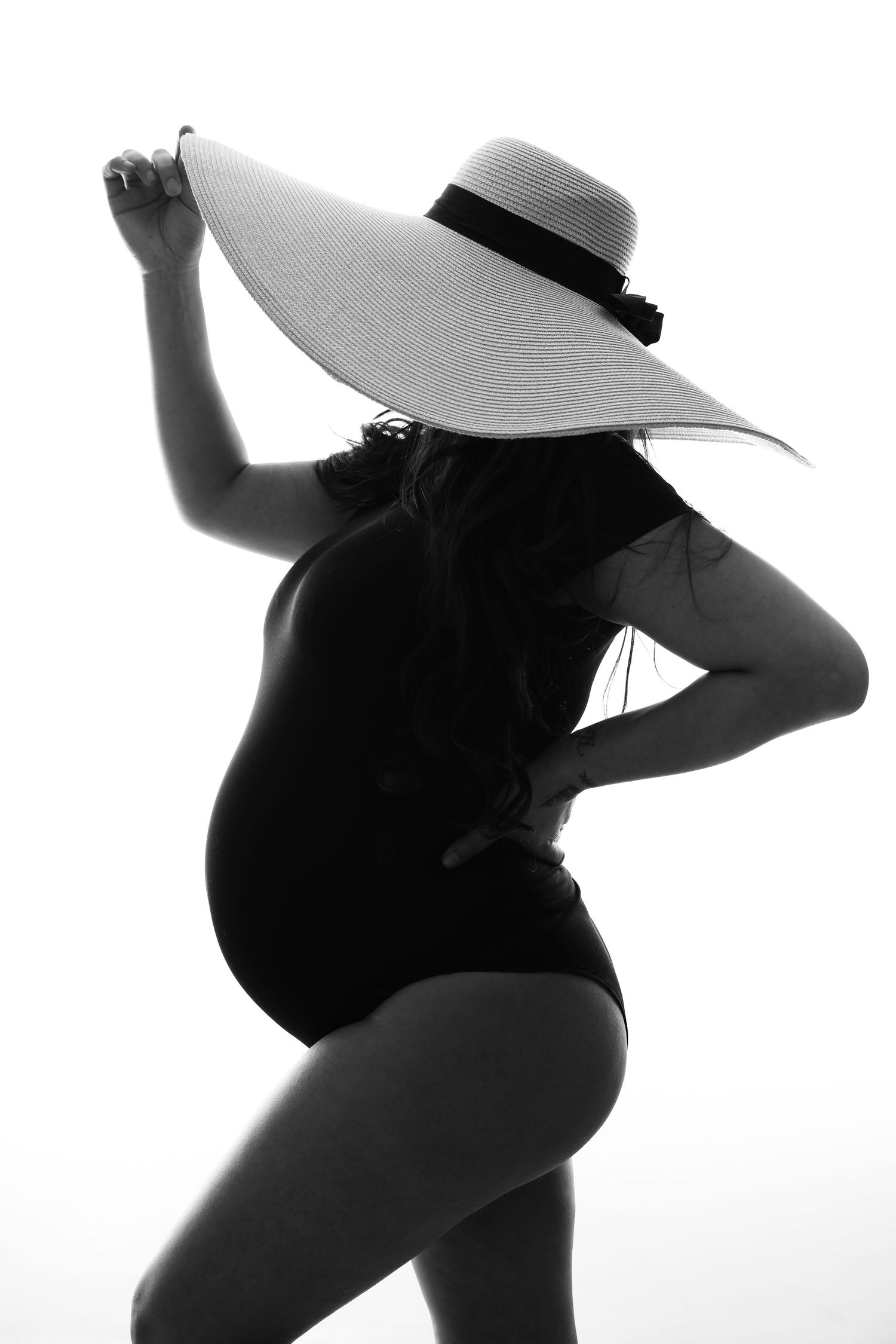 Pregnant woman silhouette in black swimsuit and wide-brim sun hat, elegant black and white maternity photo.