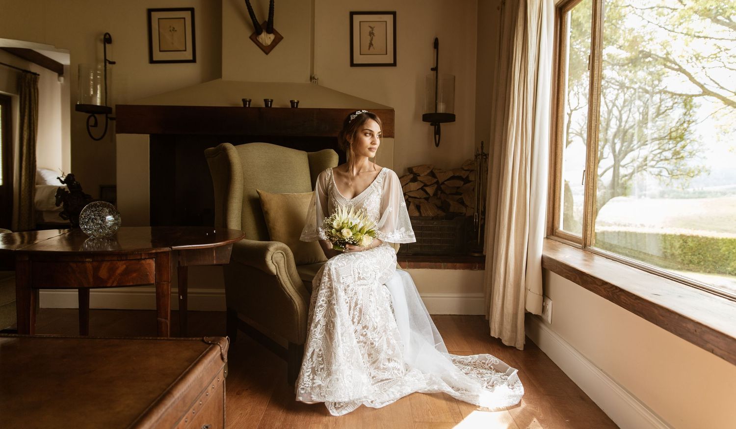 Bride in lace wedding gown sitting by window in rustic room holding bouquet.