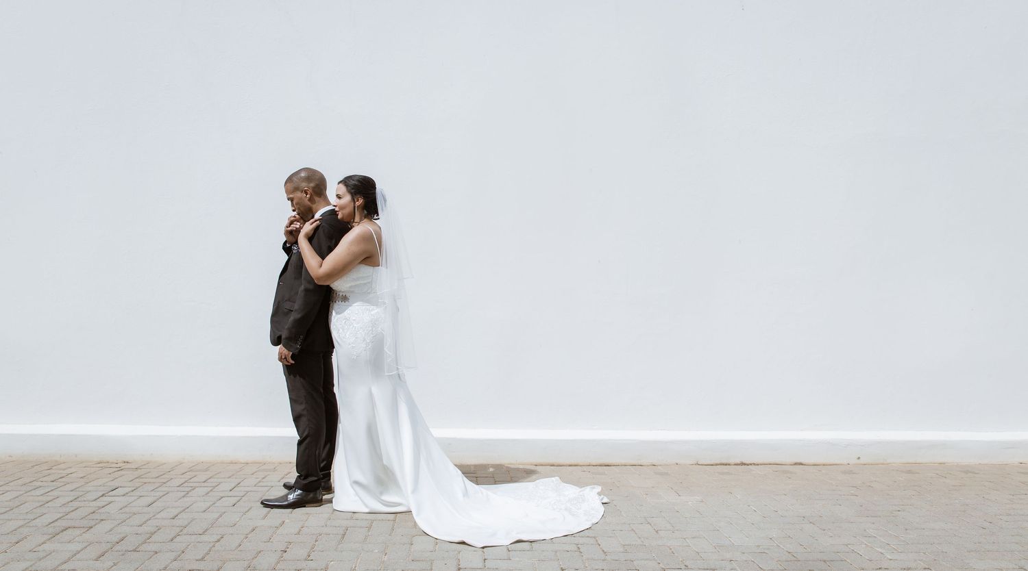 Bride and groom sharing intimate moment against white wall in wedding attire.