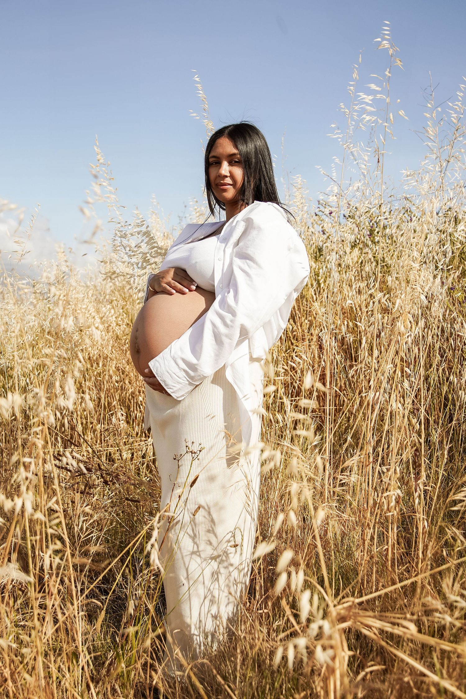 A pregnant woman in white outfit embraces her baby bump while posing in a field of tall golden wheat under blue sky.
