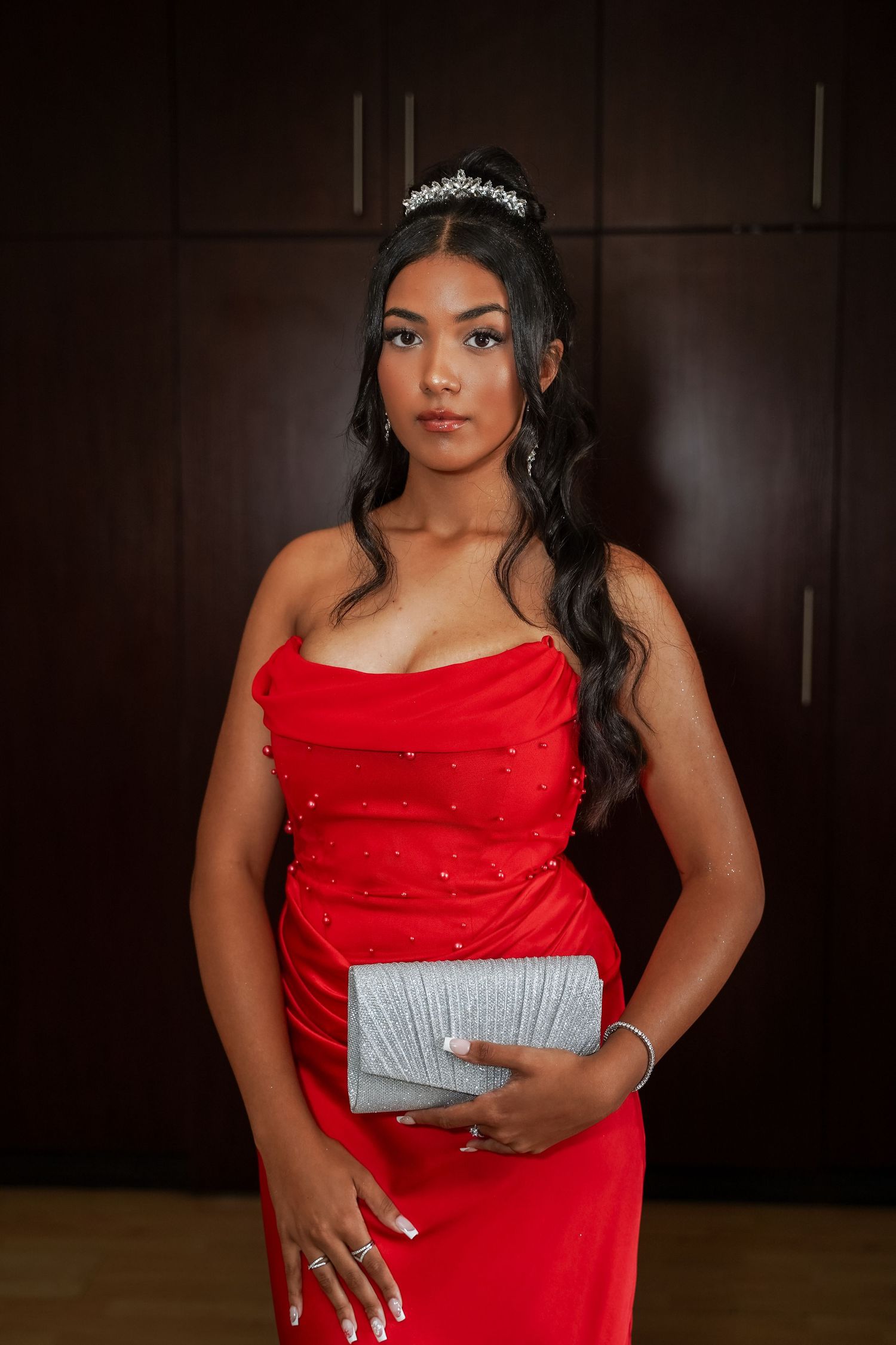 Young woman in red strapless pearl-embellished gown holds silver clutch wearing diamond tiara indoors.
