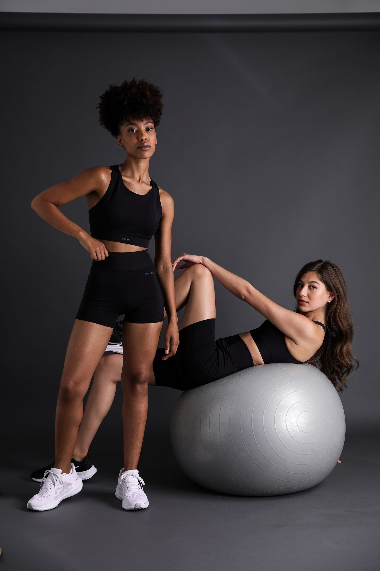 Two women in black activewear pose with a silver fitness ball against a dark gray studio background.